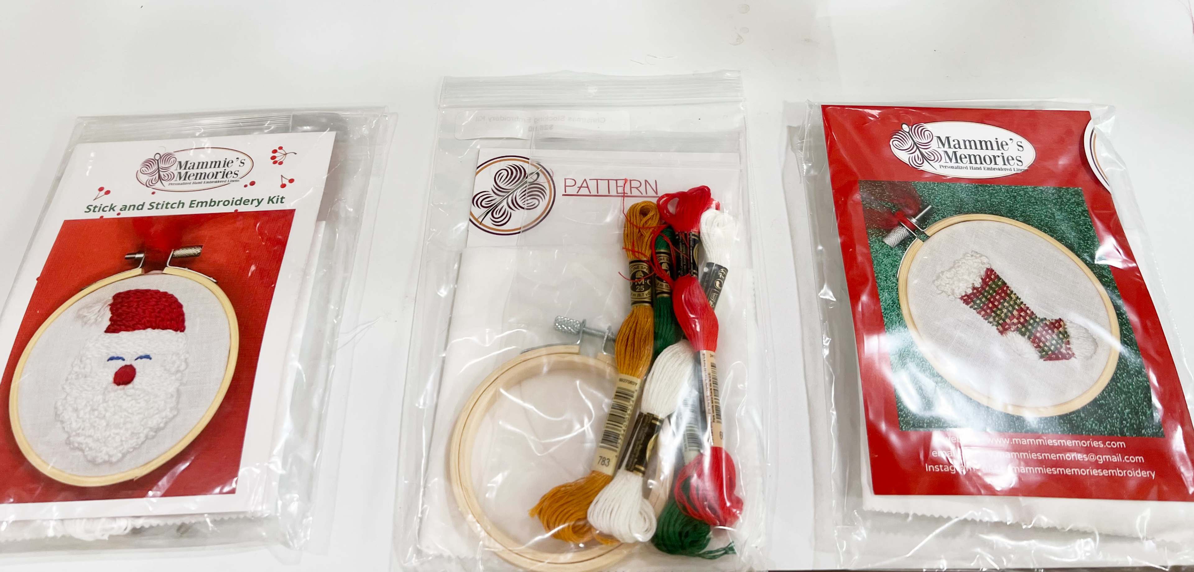 Three packaged embroidery kits featuring patterns for a Santa face and a Christmas stocking are arranged on a white background.