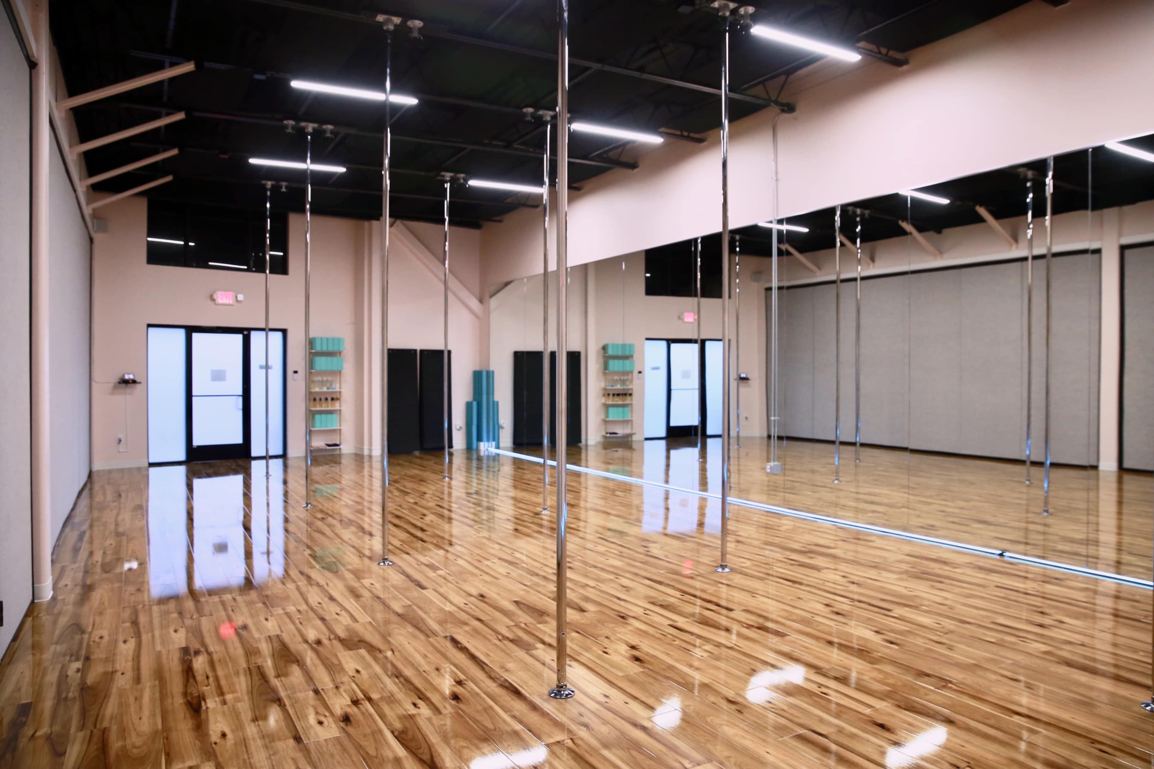 The image shows a spacious dance studio with polished wooden flooring, several reflective poles, and large mirrors along one wall.