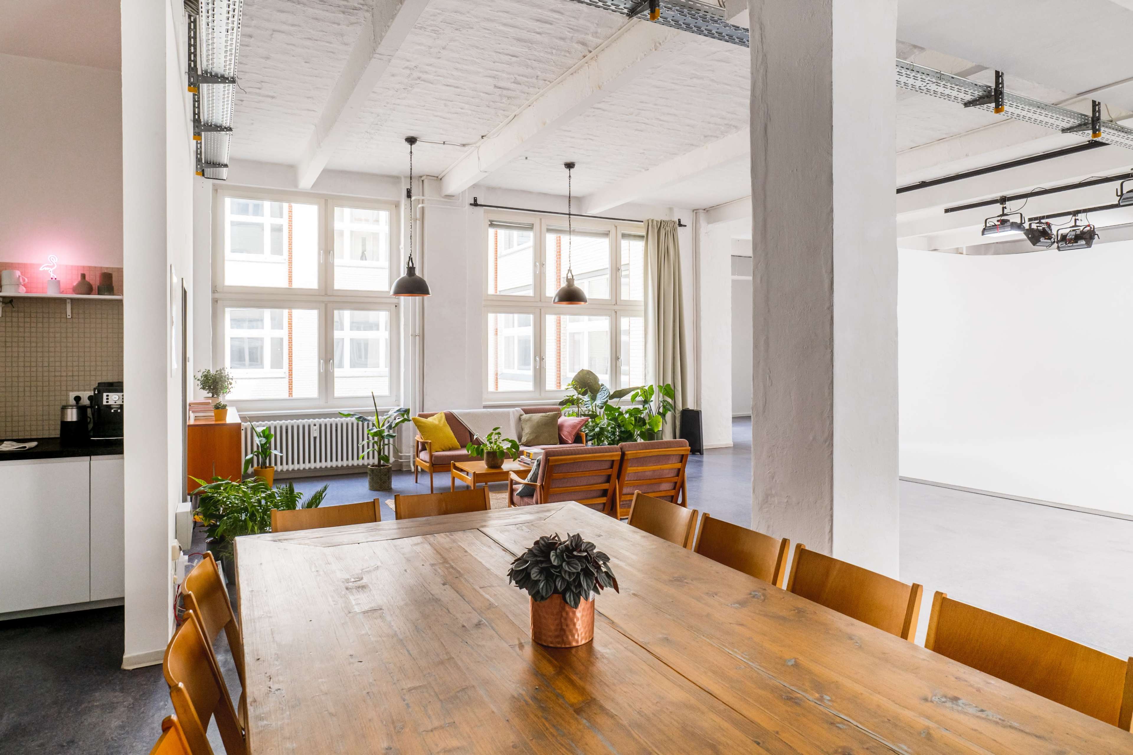 A modern interior space features a large wooden dining table surrounded by chairs, with a kitchenette on one side and a seating area filled with plants on the other.
