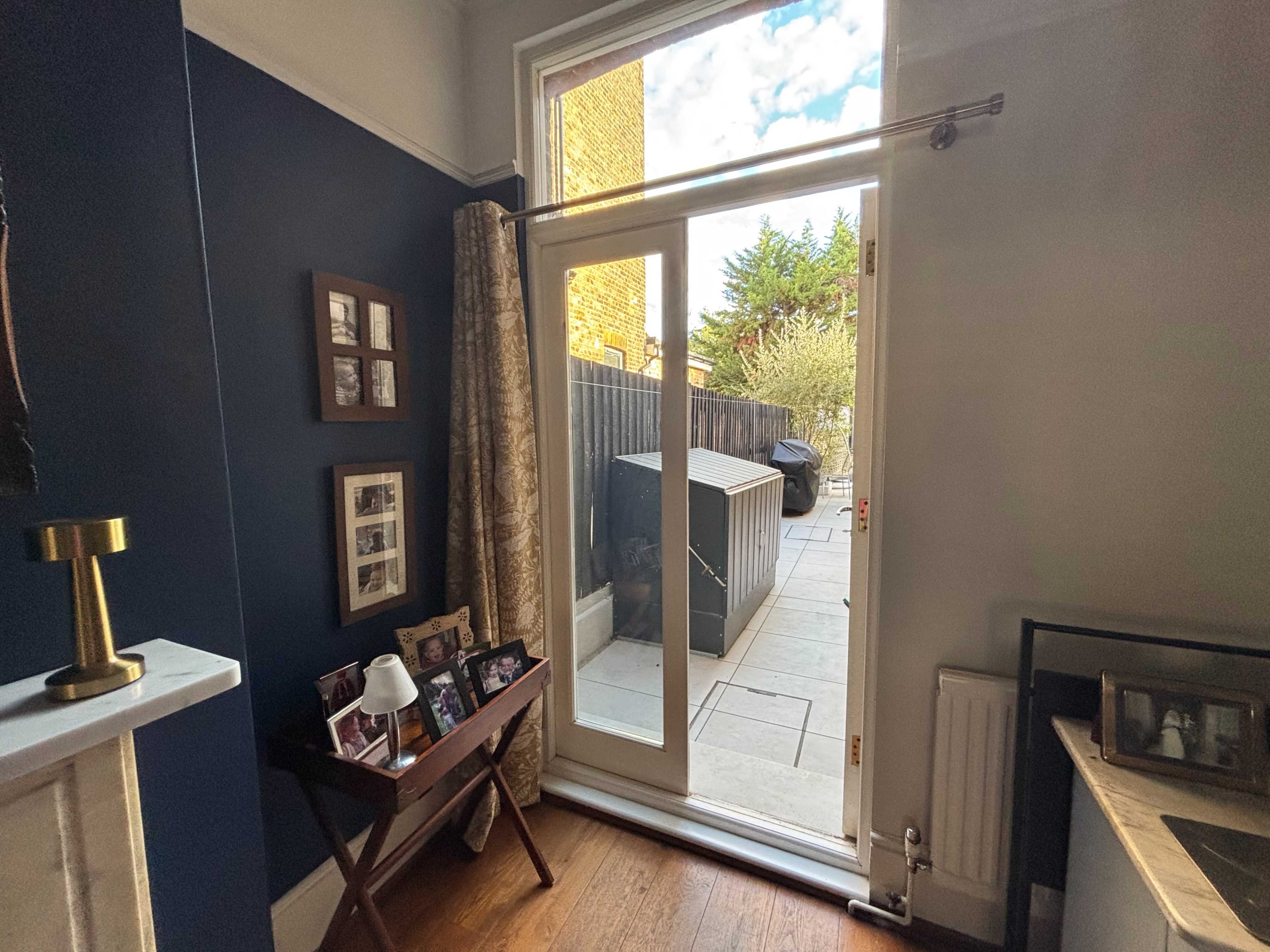 The image shows a well-lit interior space with a glass door that opens to a stone patio and garden area, framed by a dark blue wall and decorative curtains.