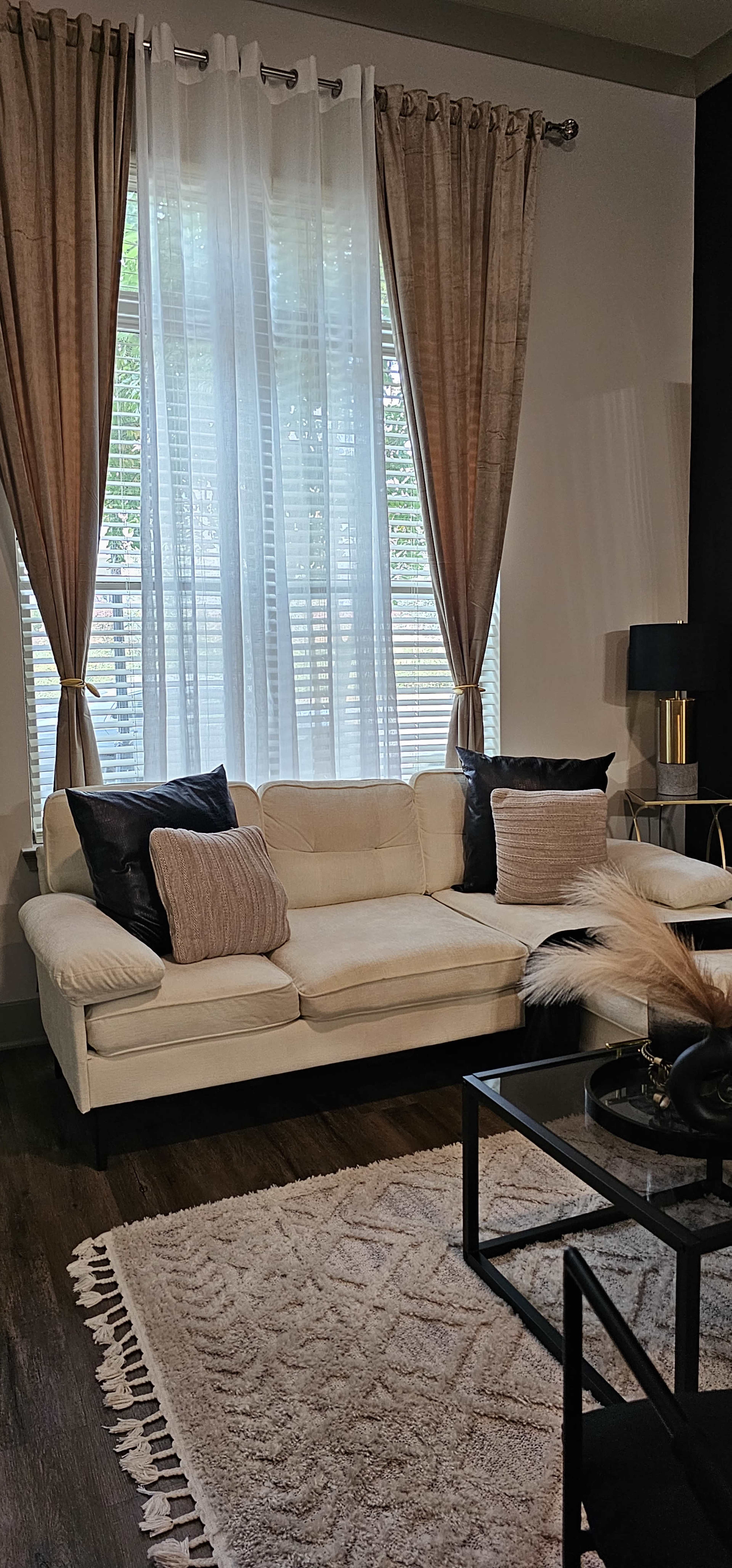 The scene features a cozy living room with a light-colored sectional sofa, decorative pillows, a glass coffee table, and large windows adorned with flowy curtains.