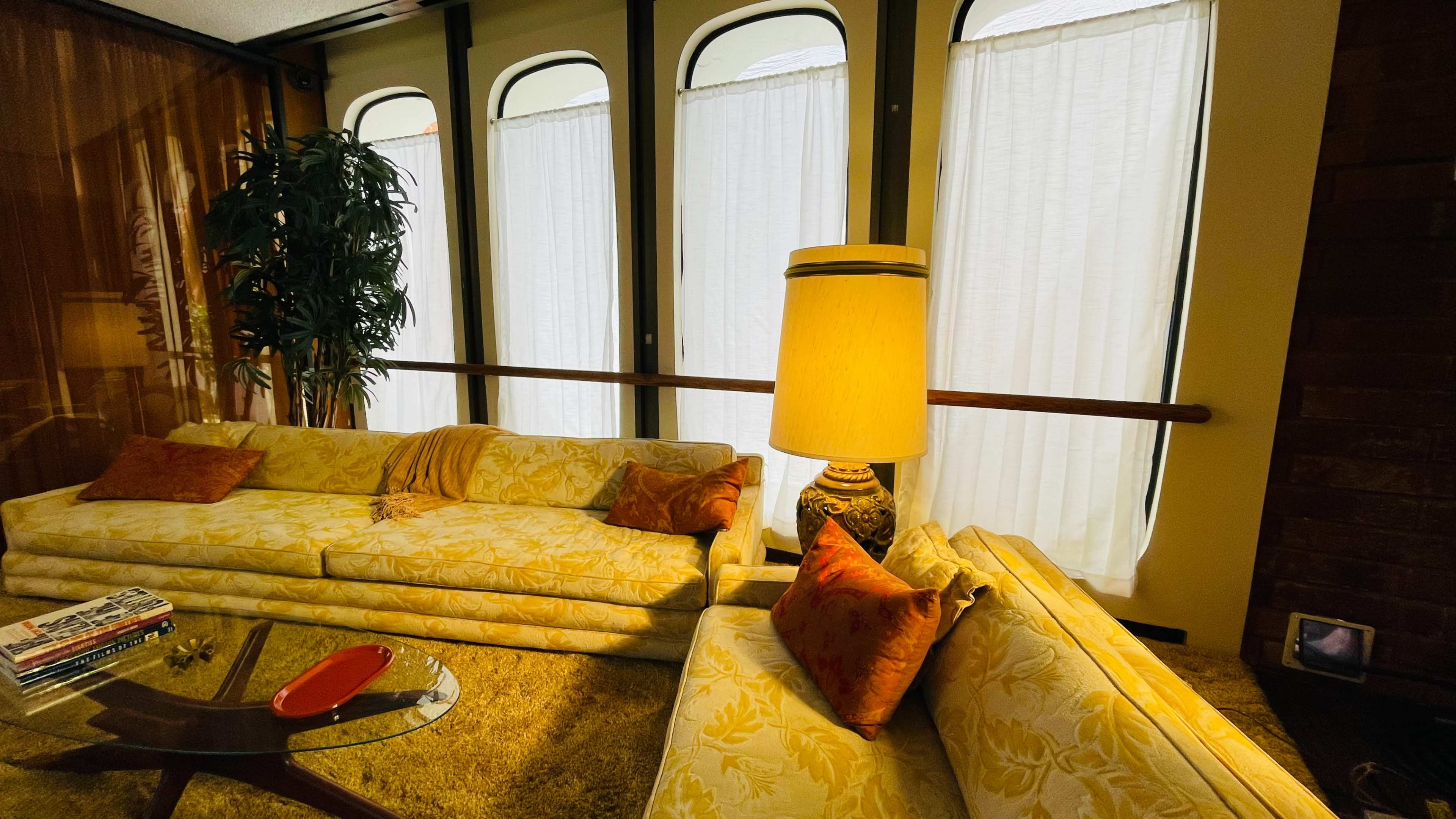 A living room features two floral-patterned sofas, a floor lamp, and large windows covered with sheer white curtains.