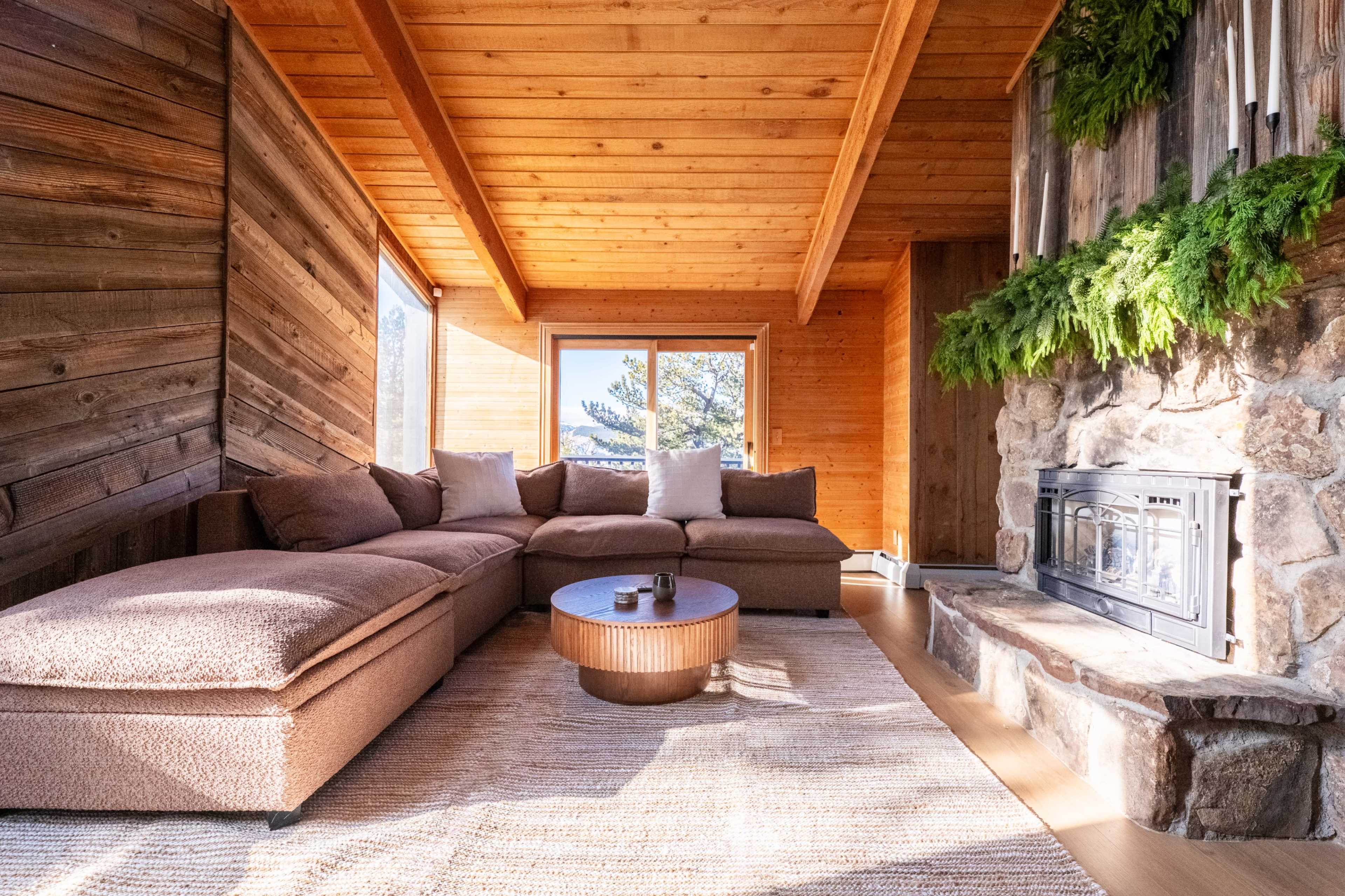 The image shows a cozy living room with a wooden ceiling, a large sectional sofa, a round coffee table, and a stone fireplace, enhanced by natural light from the windows.