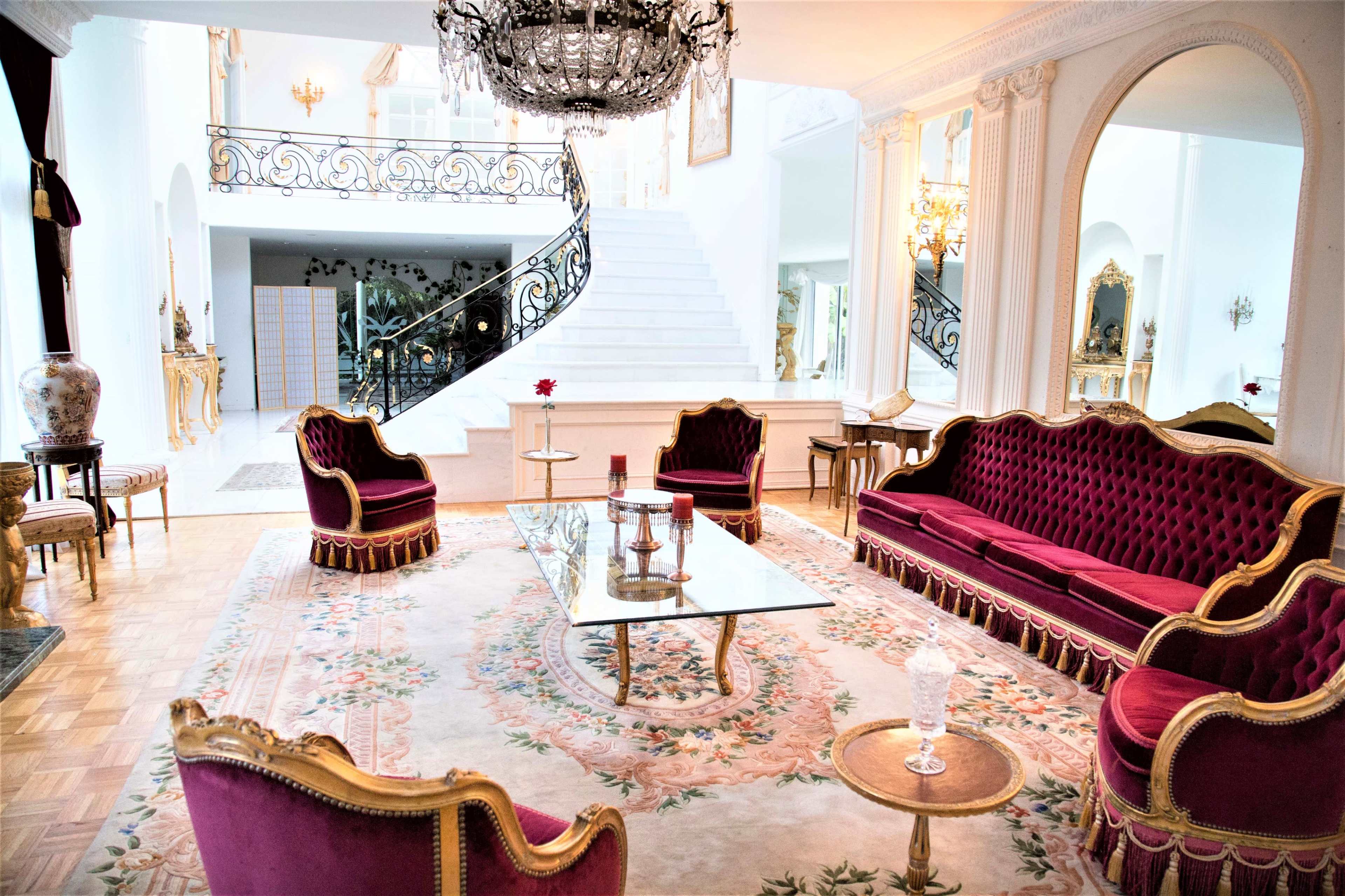 An elegant living room featuring ornate red seating, a glass coffee table, and a grand staircase in the background.