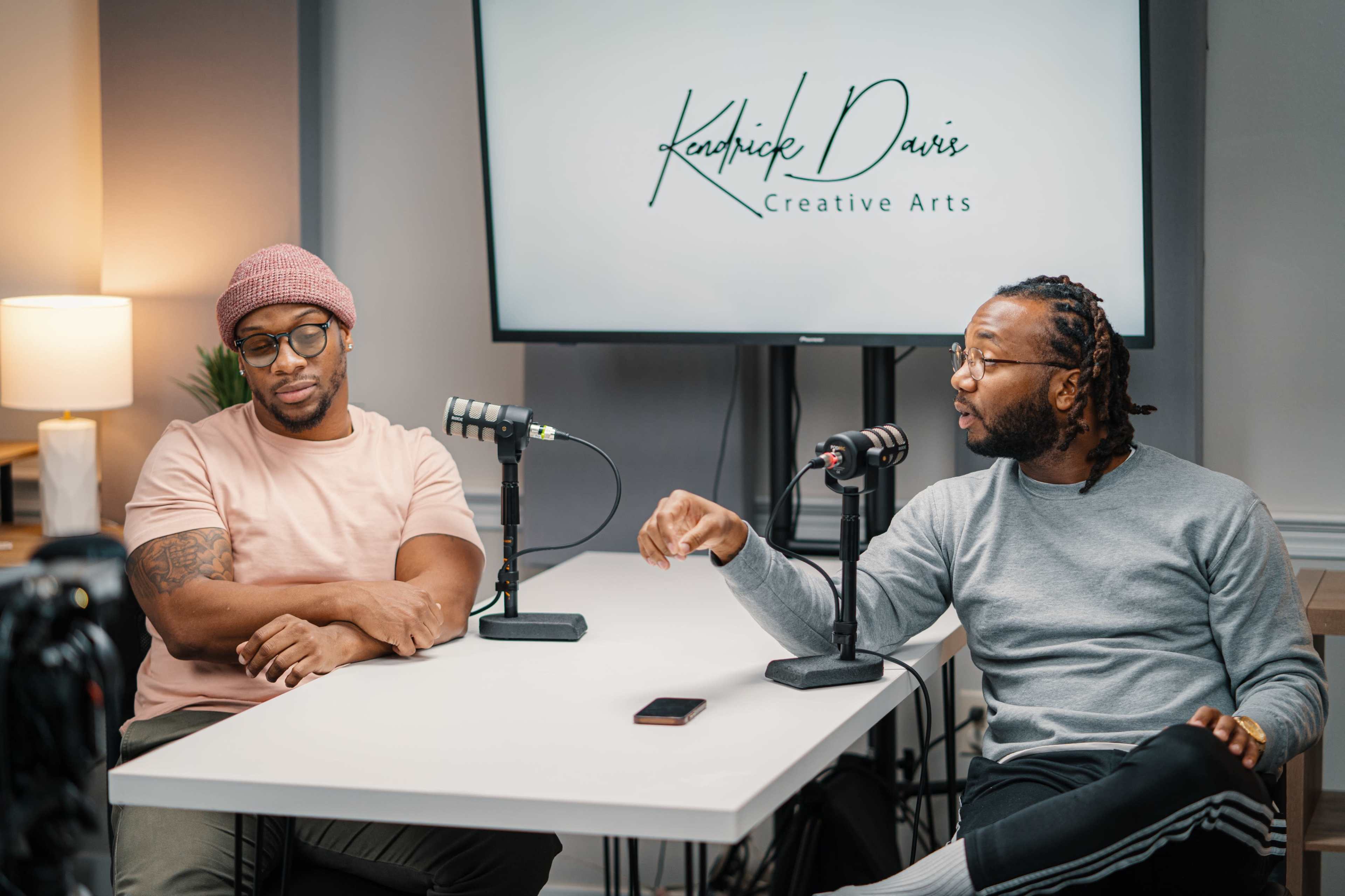 Two men are seated at a table with microphones in front of them, discussing in a well-lit room featuring a display screen that reads "Kendrick Davis Creative Arts."