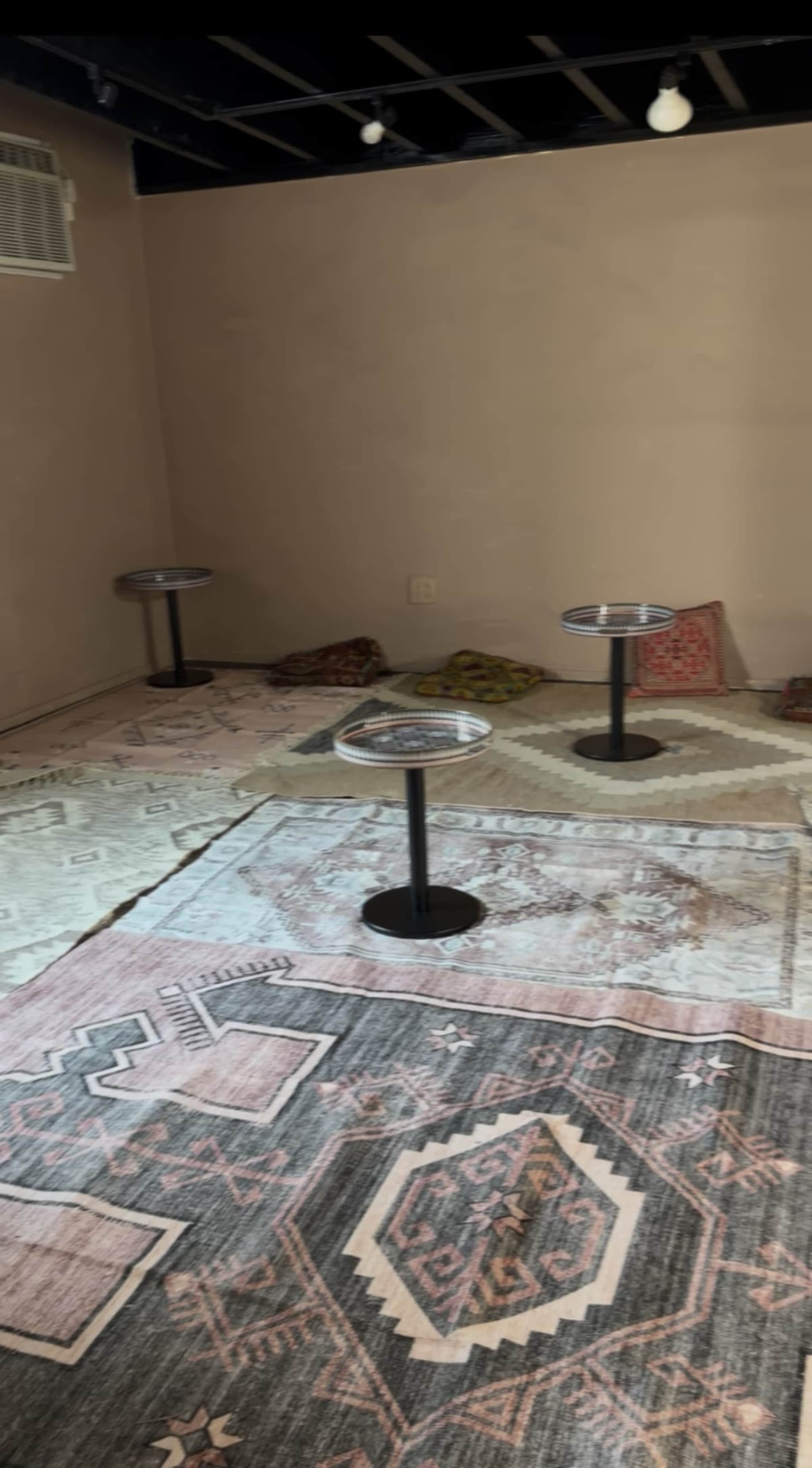 The image shows a sparsely furnished room with a few round tables on stands and decorative rugs on the floor.