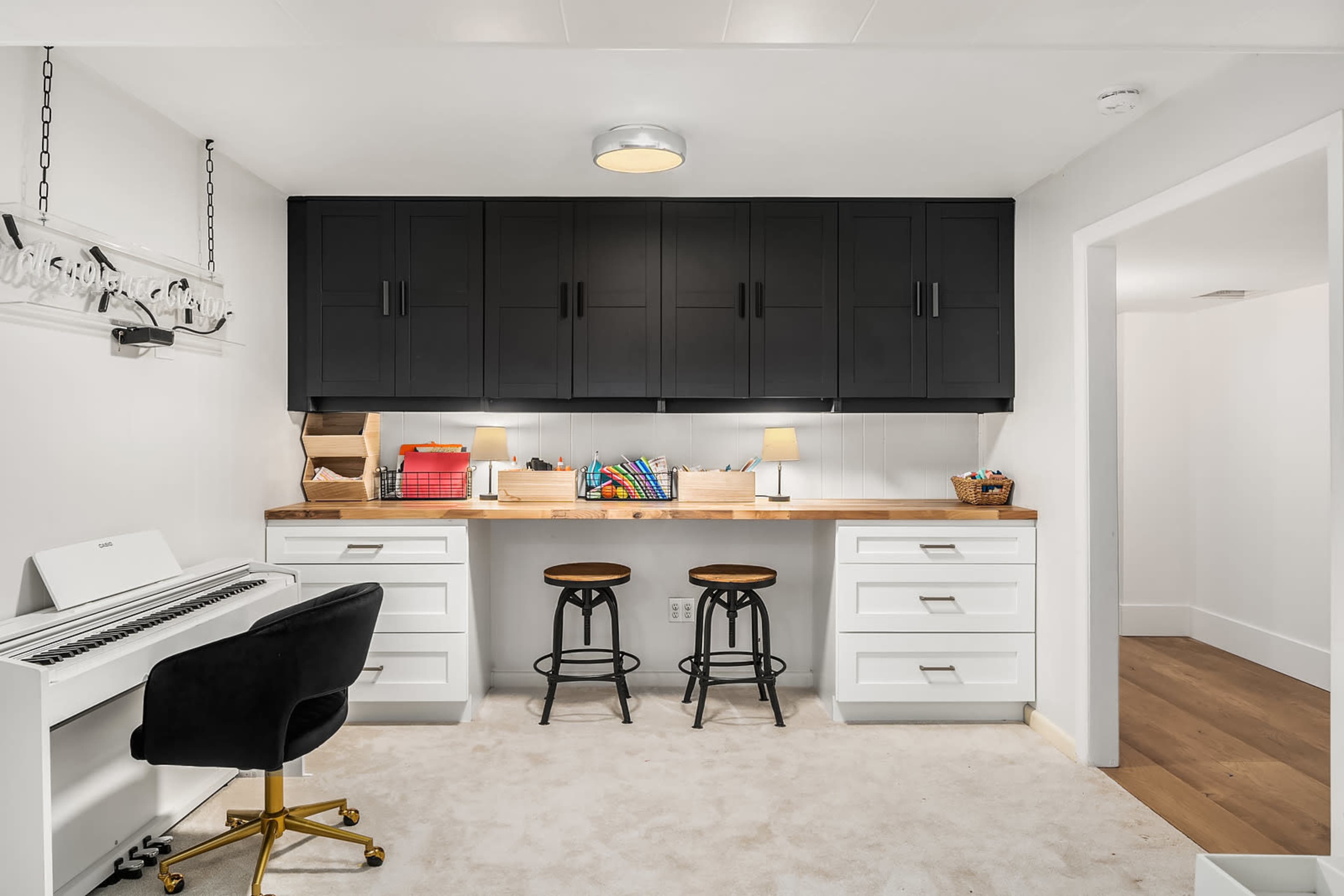 The room features a workspace with a white piano, two black stools, and a dark cabinet above a wooden countertop.