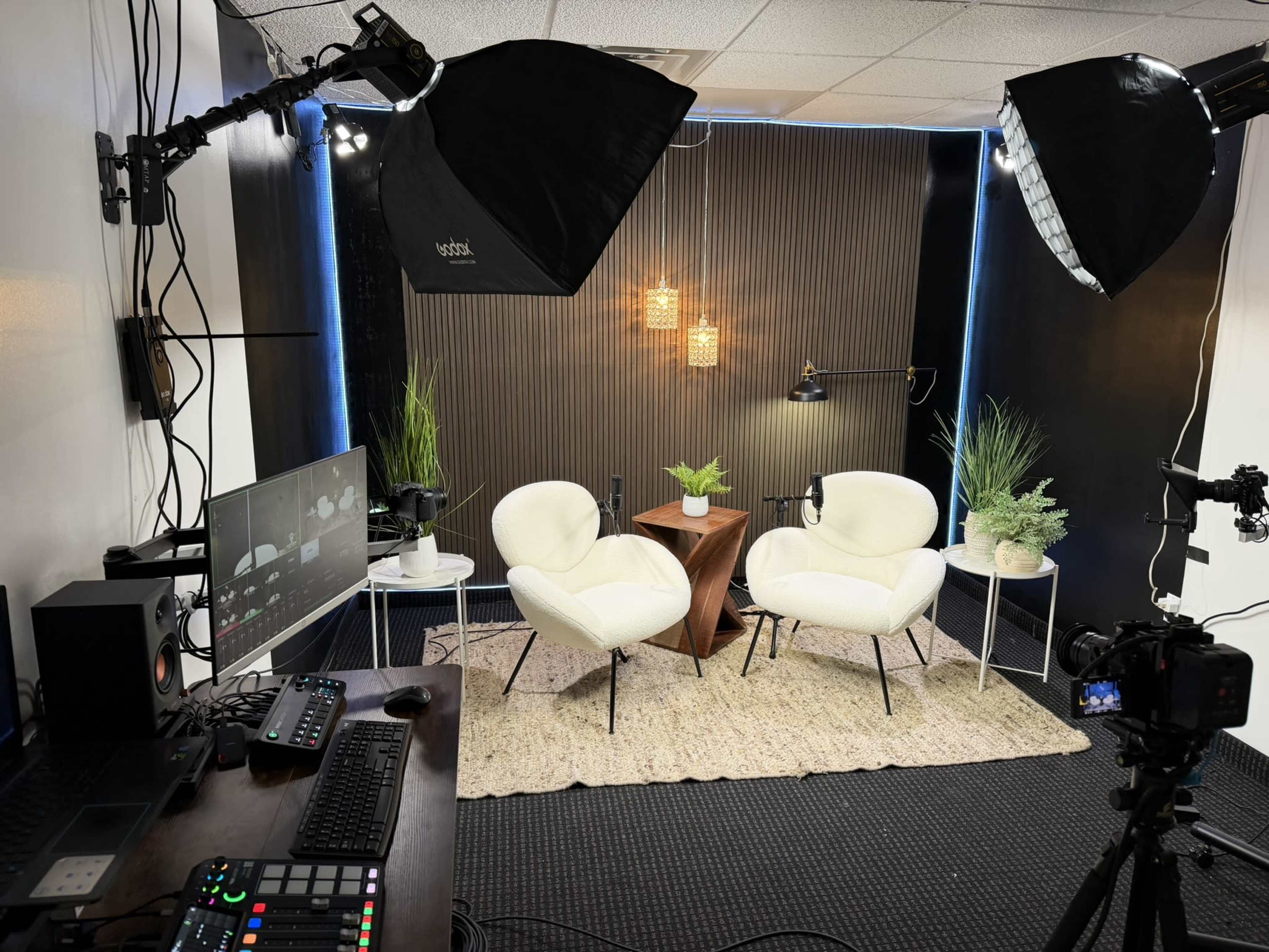 The image shows a modern, well-lit studio setup featuring two white armchairs, a wooden table, and decorative plants against a backdrop with vertical slats.