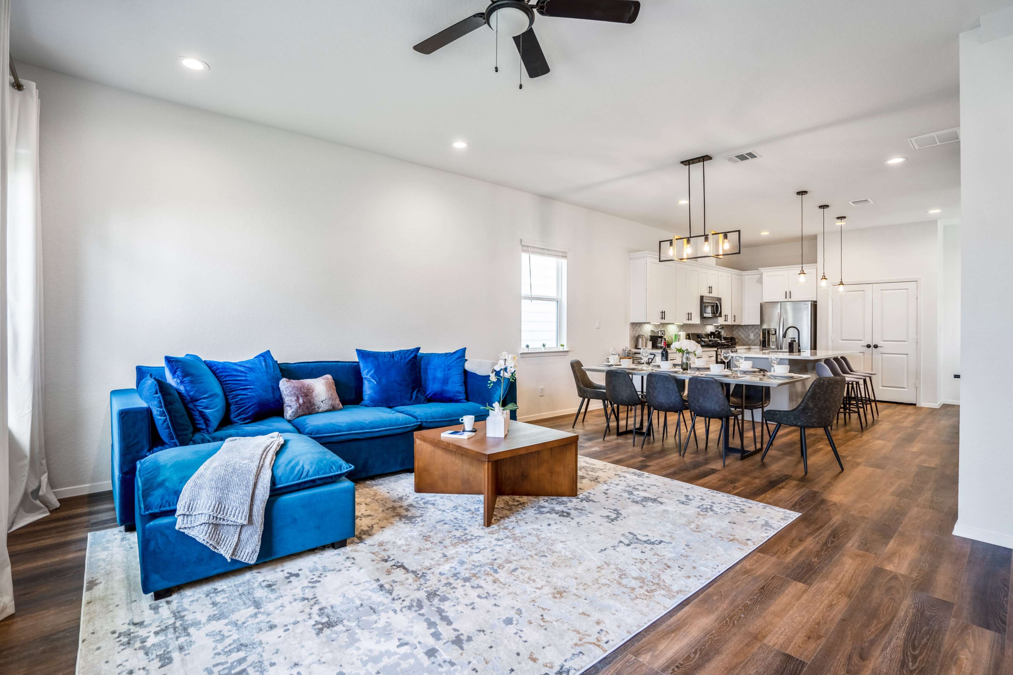 A modern living room features a blue sectional sofa, a coffee table, and a dining area with a table set for six in an open-concept space.