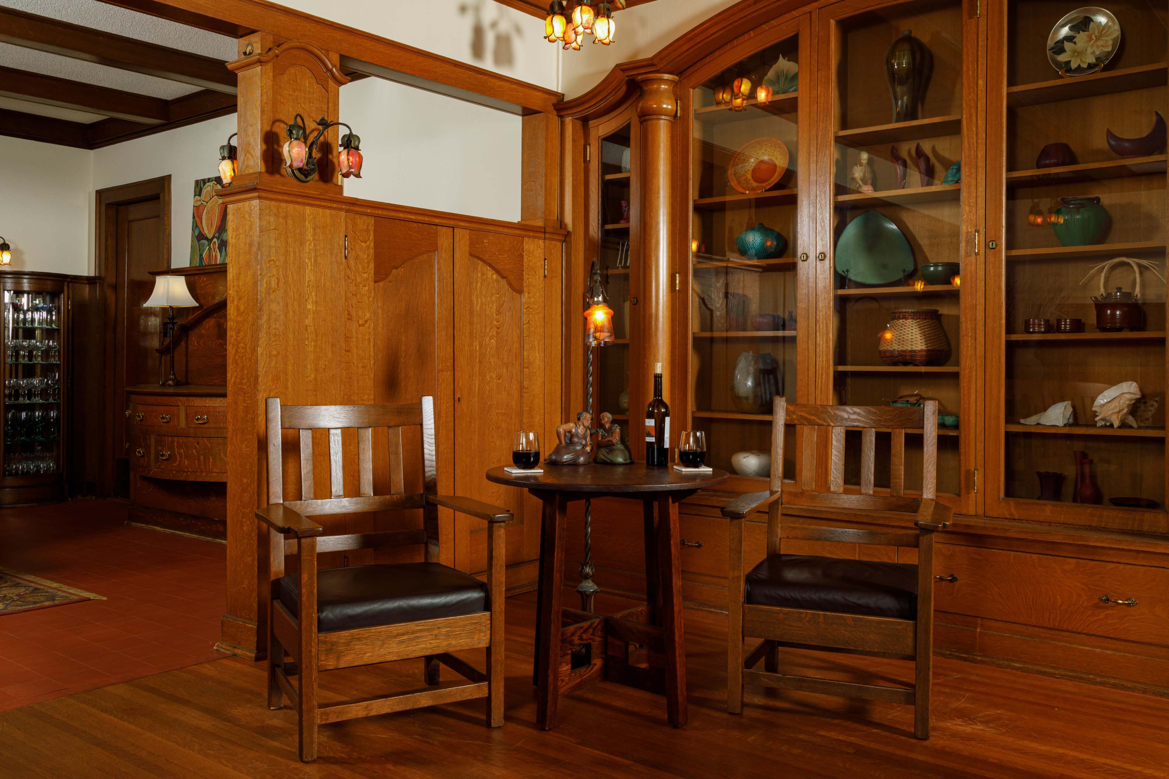 A cozy interior featuring two wooden chairs positioned around a small table, with a wine bottle and glasses on it, alongside a large wooden cabinet displaying various decorative items.