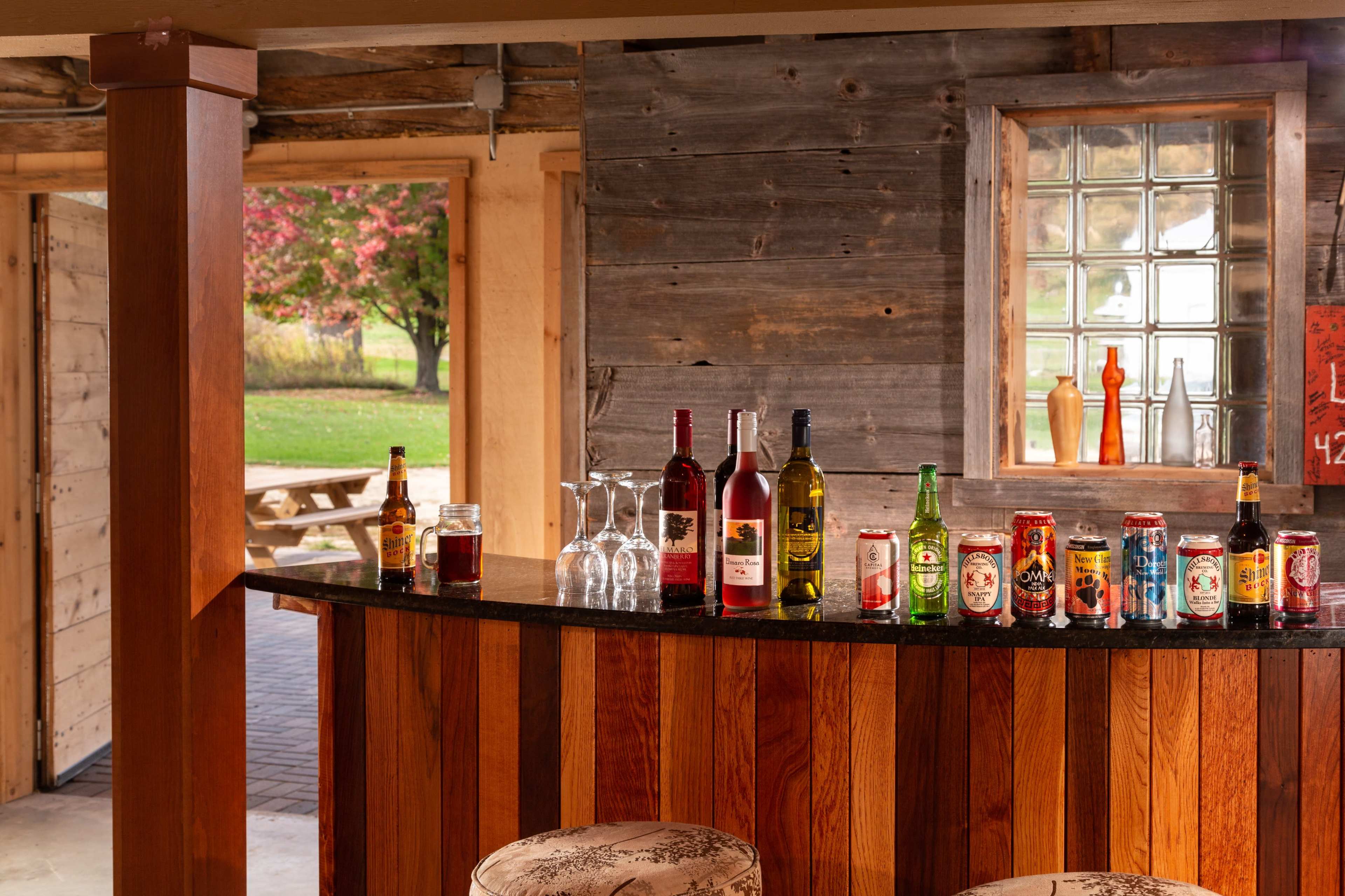 A wooden bar displays a variety of alcoholic beverages and glasses against a backdrop of a rustic interior and a window with a view of the outdoors.