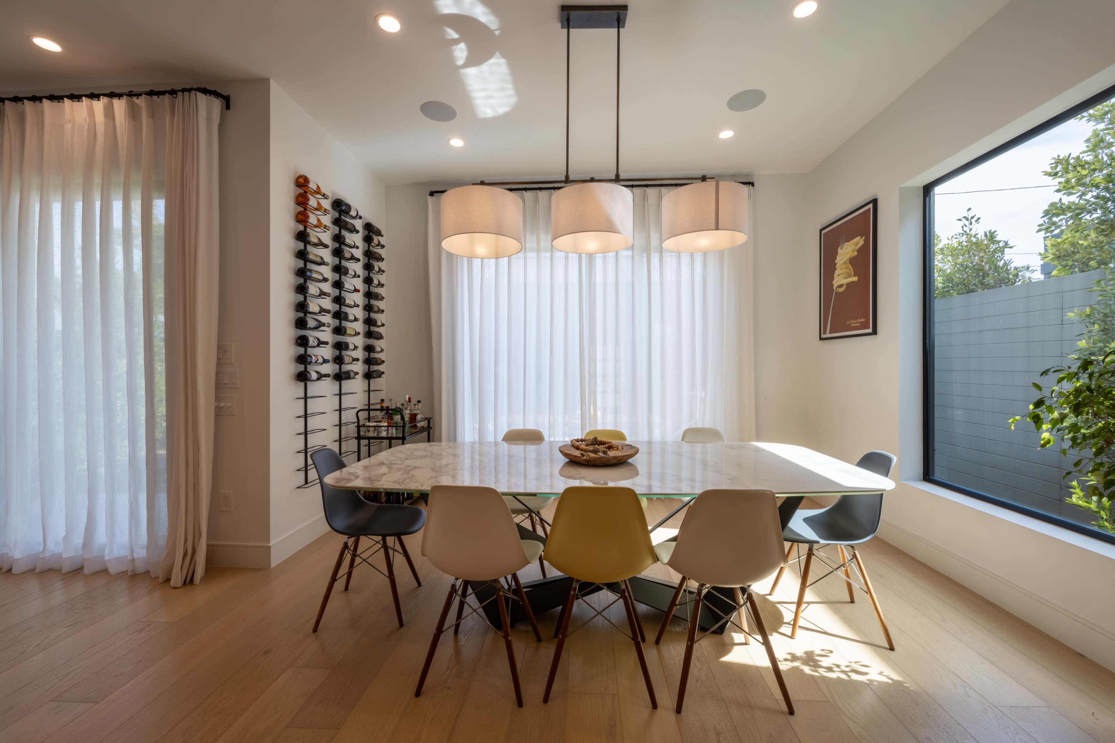 A modern dining room features a large marble table surrounded by colorful chairs, with a wine rack on one wall and large windows allowing natural light to filter through sheer curtains.