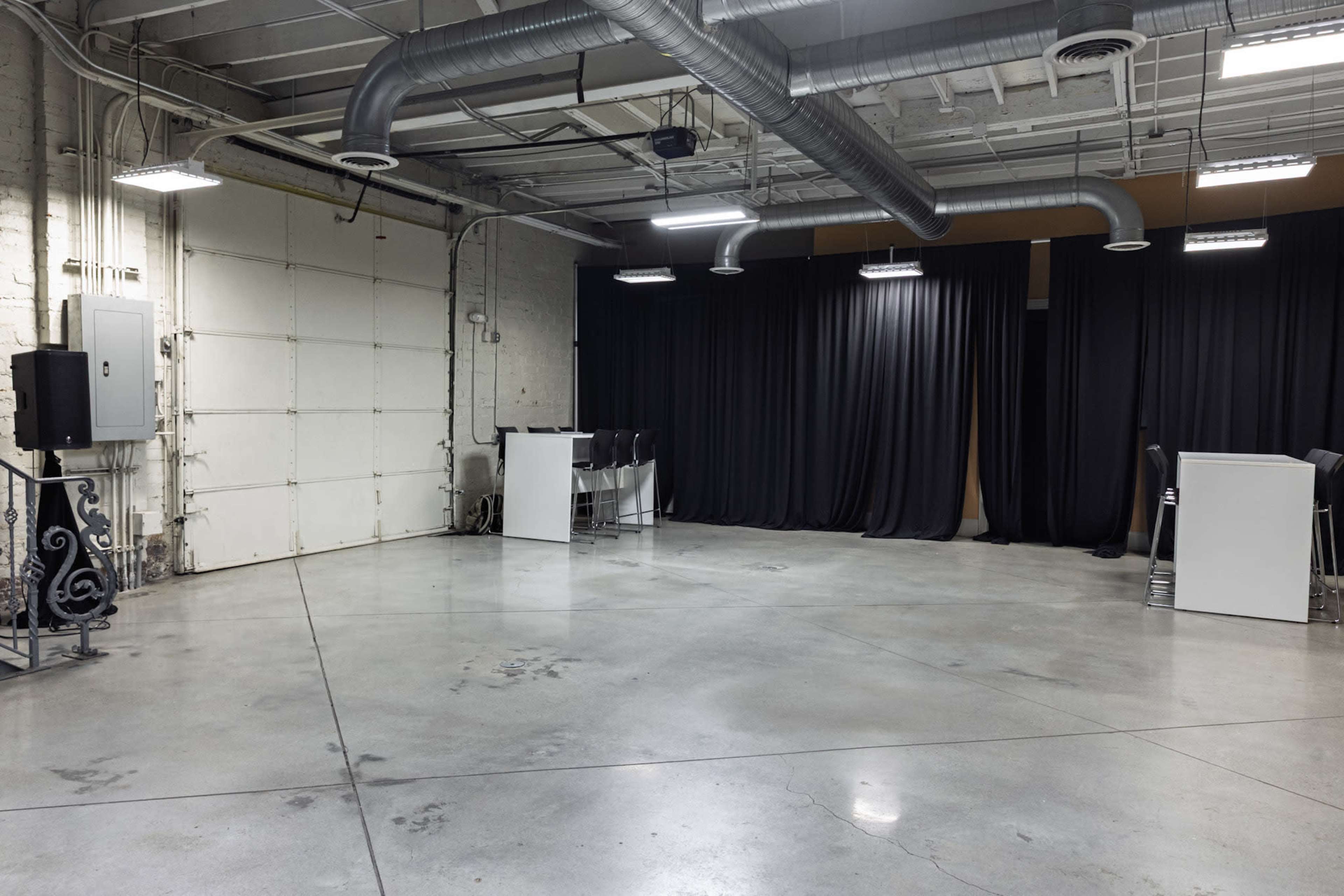 The image shows an empty industrial space with concrete flooring, exposed ventilation, and black curtains covering an area on the right.