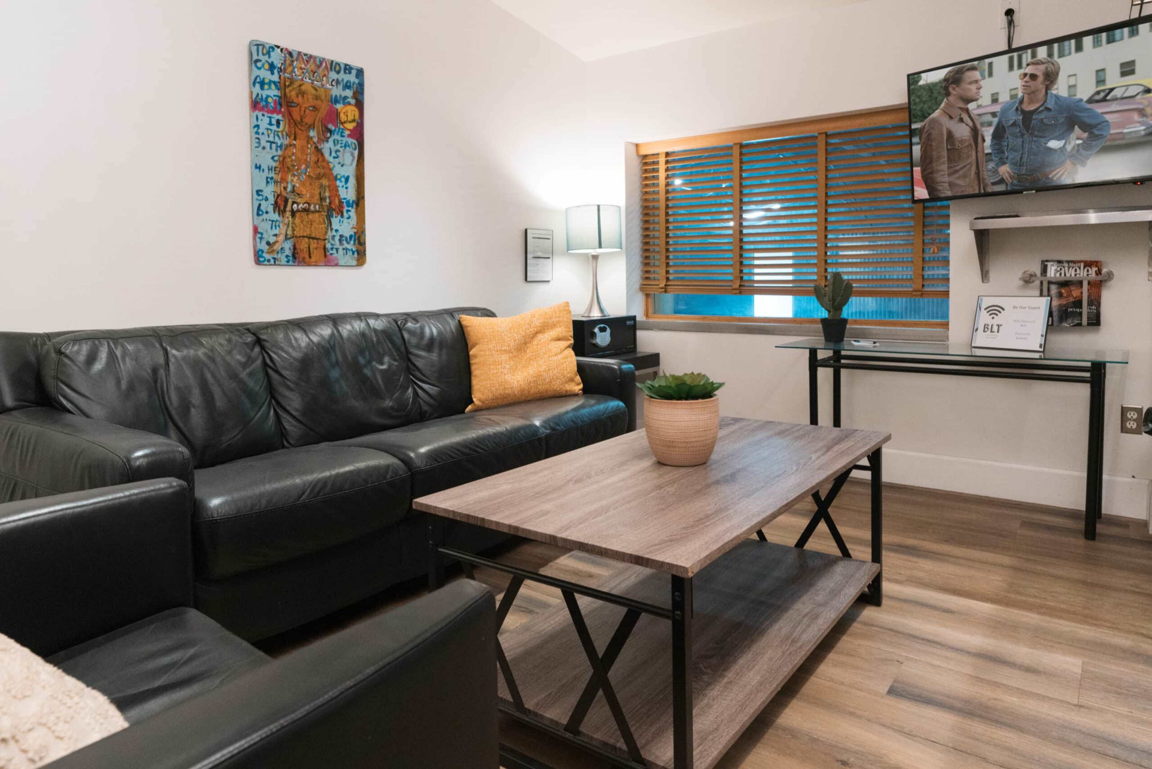 The image shows a modern living room with a black leather sofa, a wooden coffee table, and a television mounted on the wall.