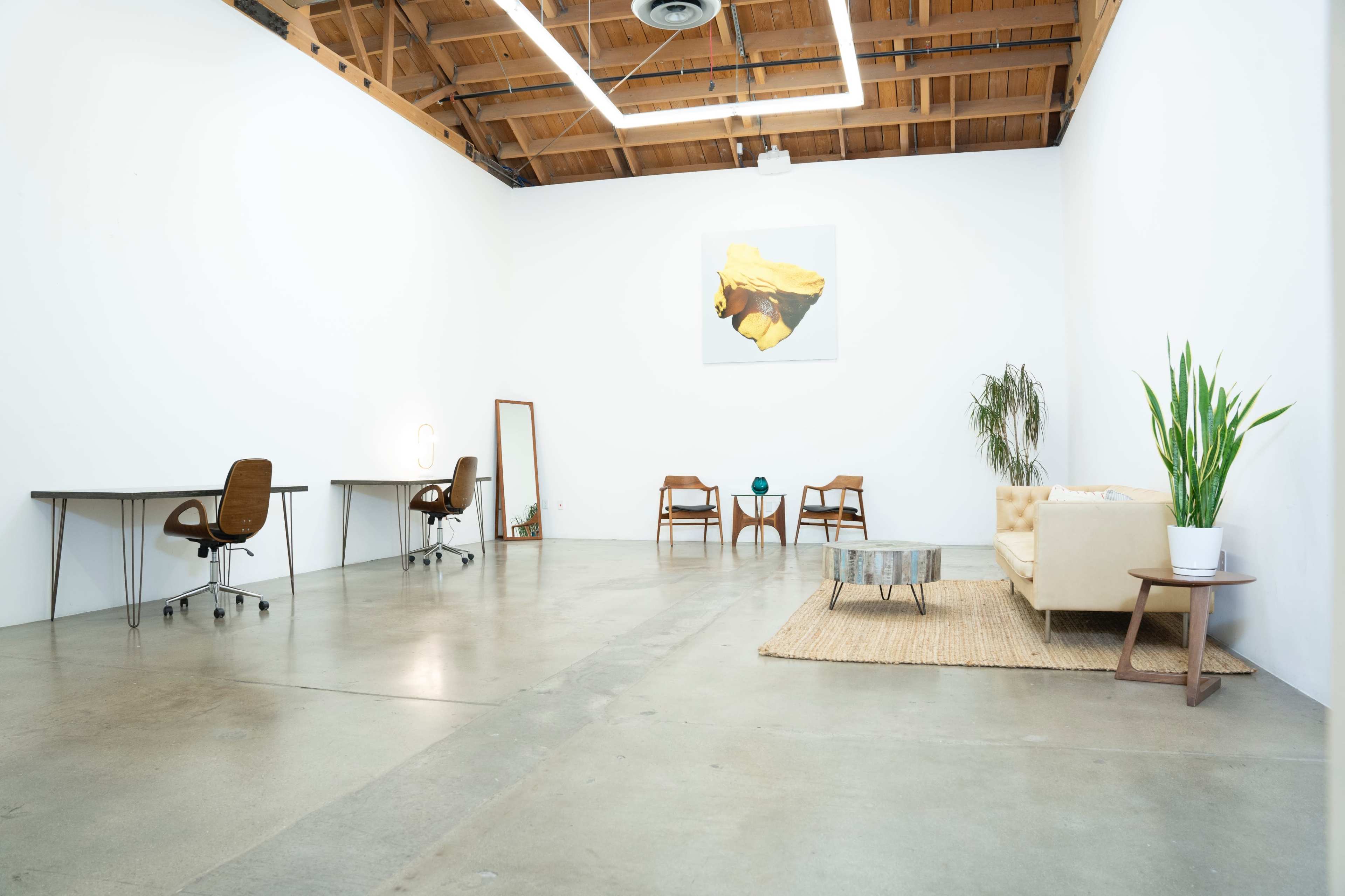 A spacious, minimalist office with two desks, a chair, a couch, and potted plants under wooden beams and a white ceiling.