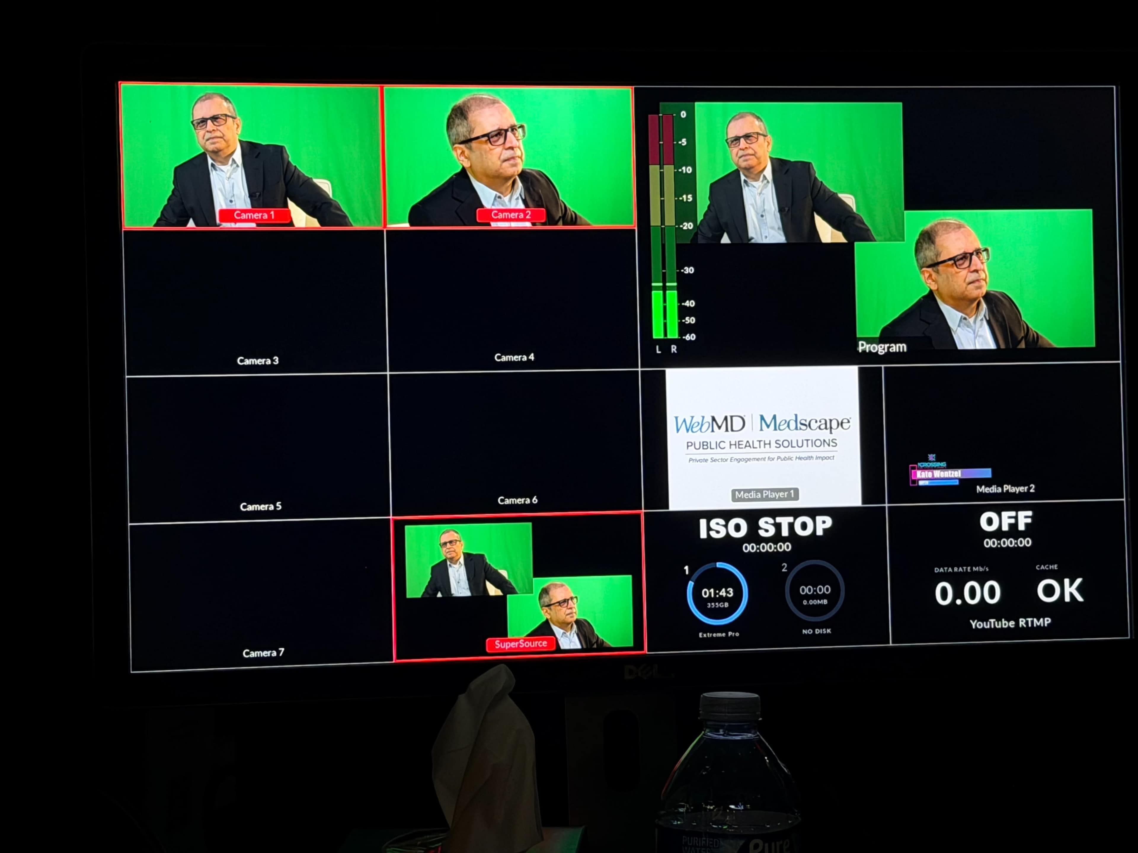The image shows a video monitoring screen with multiple camera feeds displaying a man in glasses seated in front of a green screen.