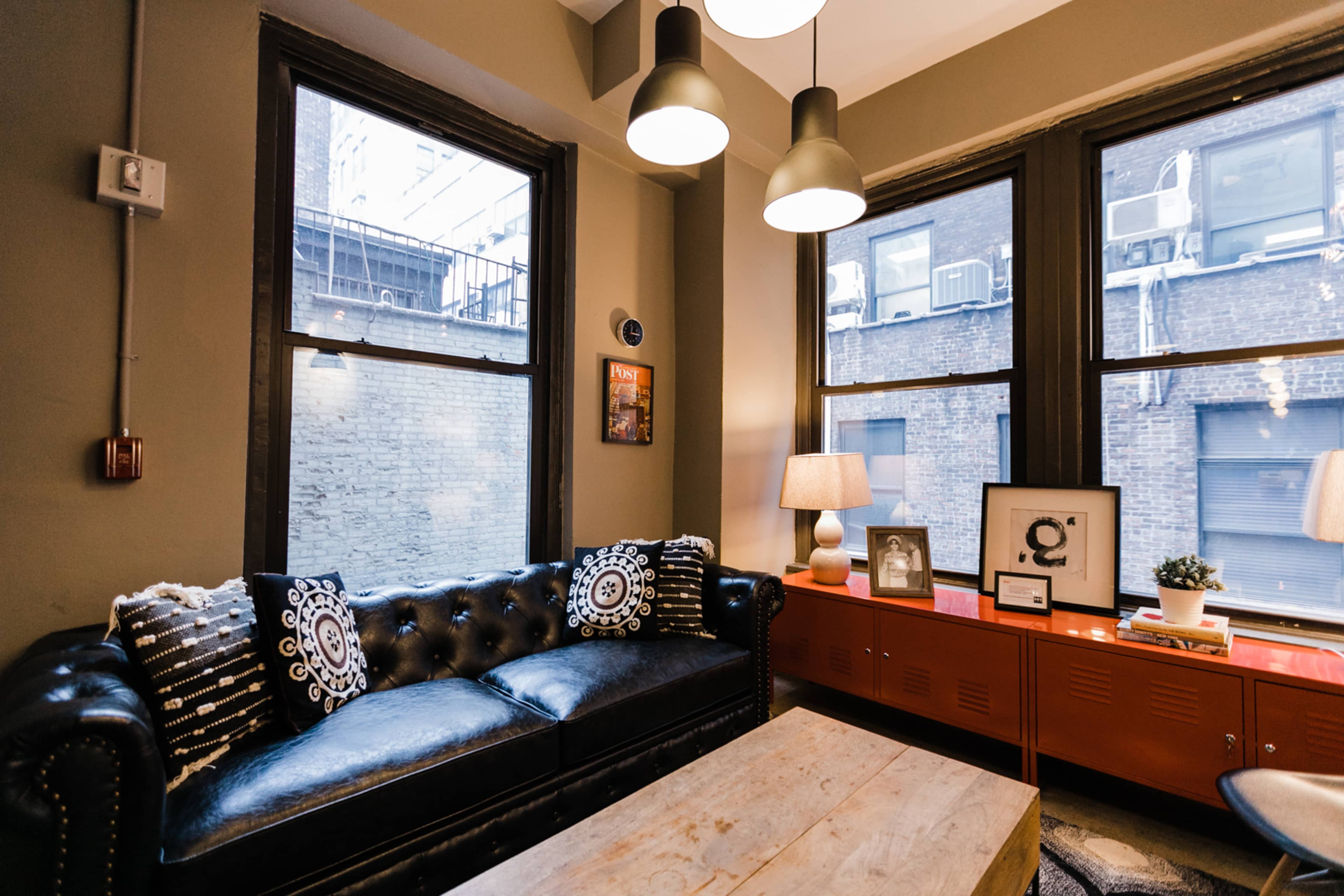 A leather sofa with decorative pillows is positioned in front of large windows, illuminating a room with a wooden coffee table and a series of lamps.