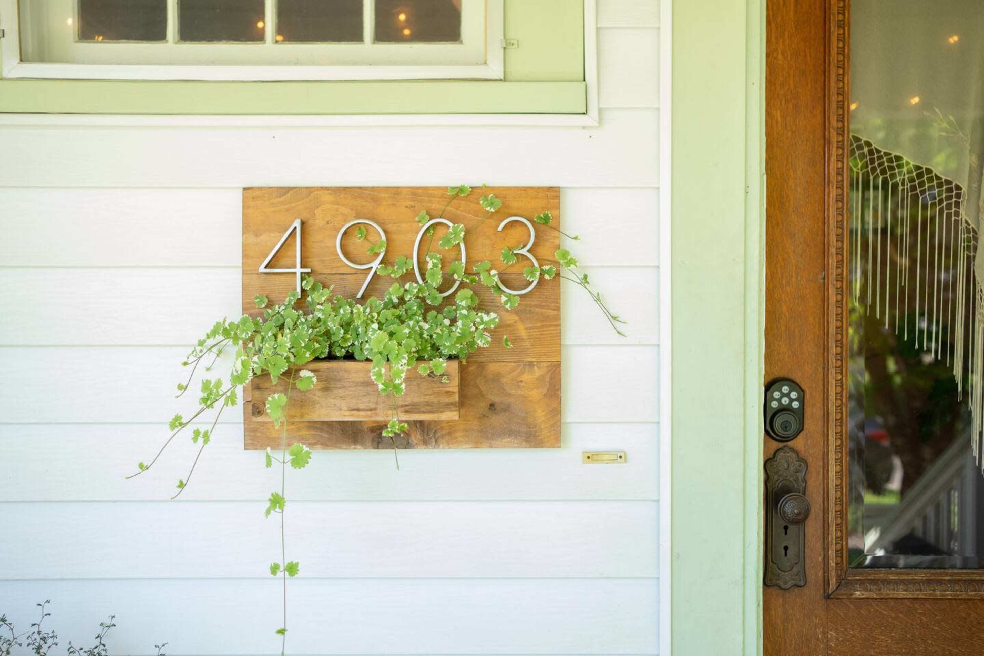A wooden mailbox holder with the number "4963" and a planter filled with green foliage is mounted on a white wall beside a door with ornate detailing.
