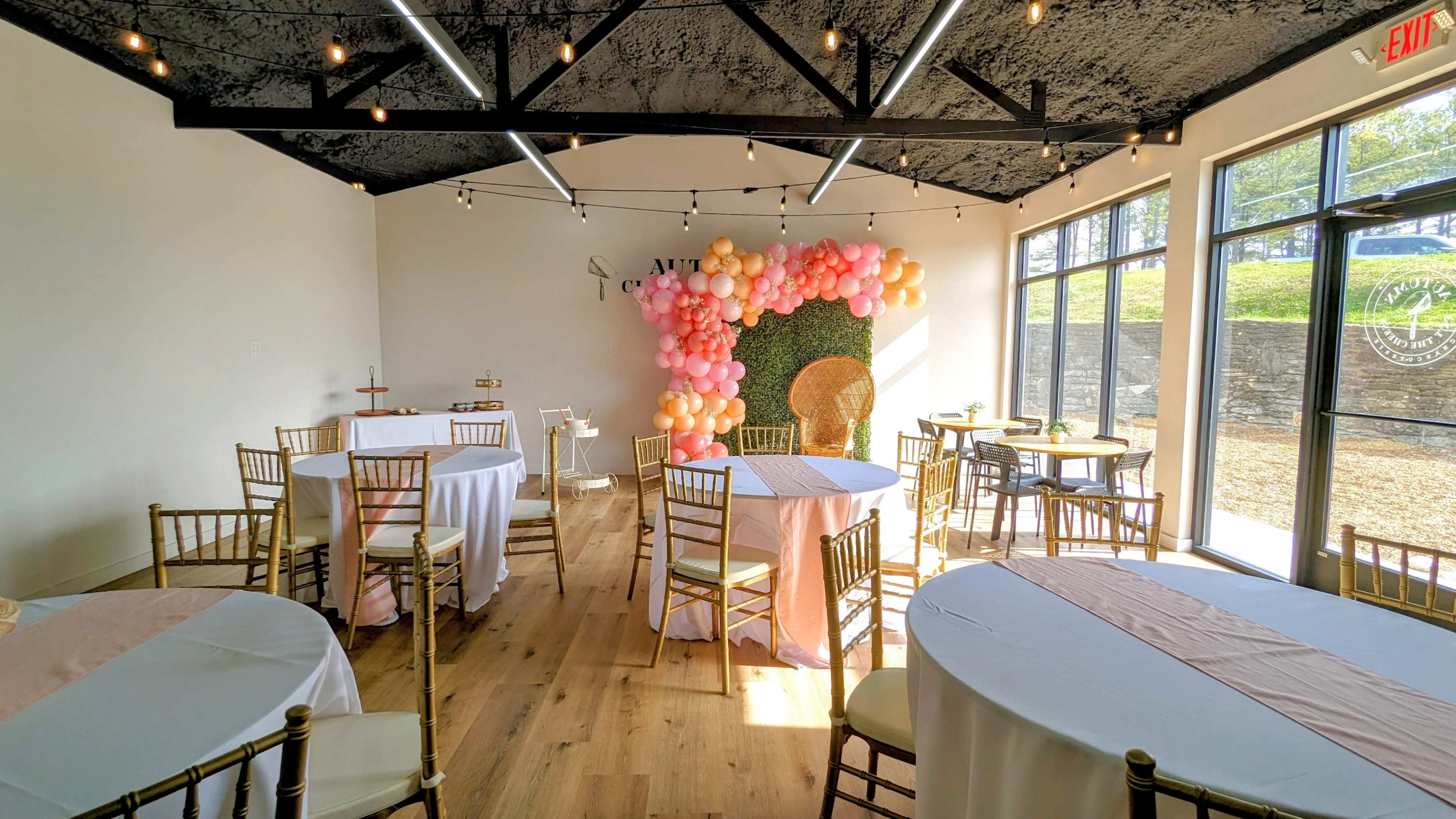 A well-lit event space with round tables set up, decorated with pastel balloons on a wall, and golden chairs arranged around the tables.