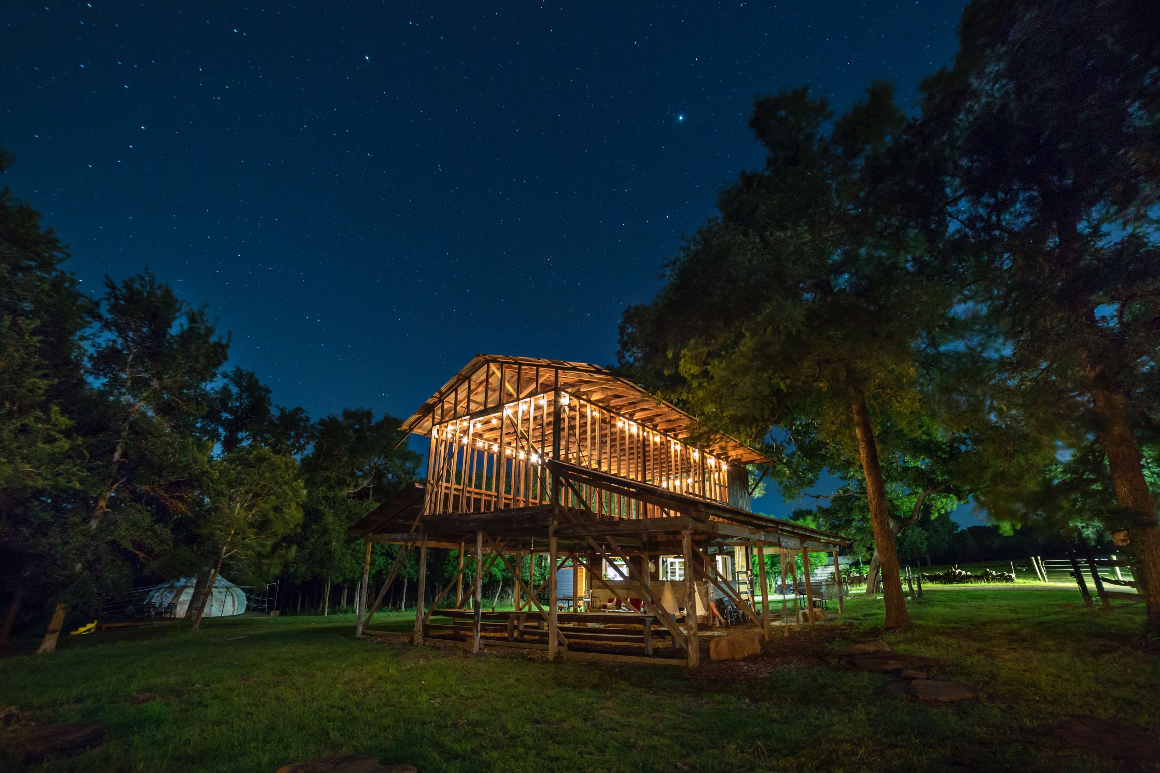 A partially constructed wooden structure is illuminated by string lights under a starry night sky, surrounded by trees.