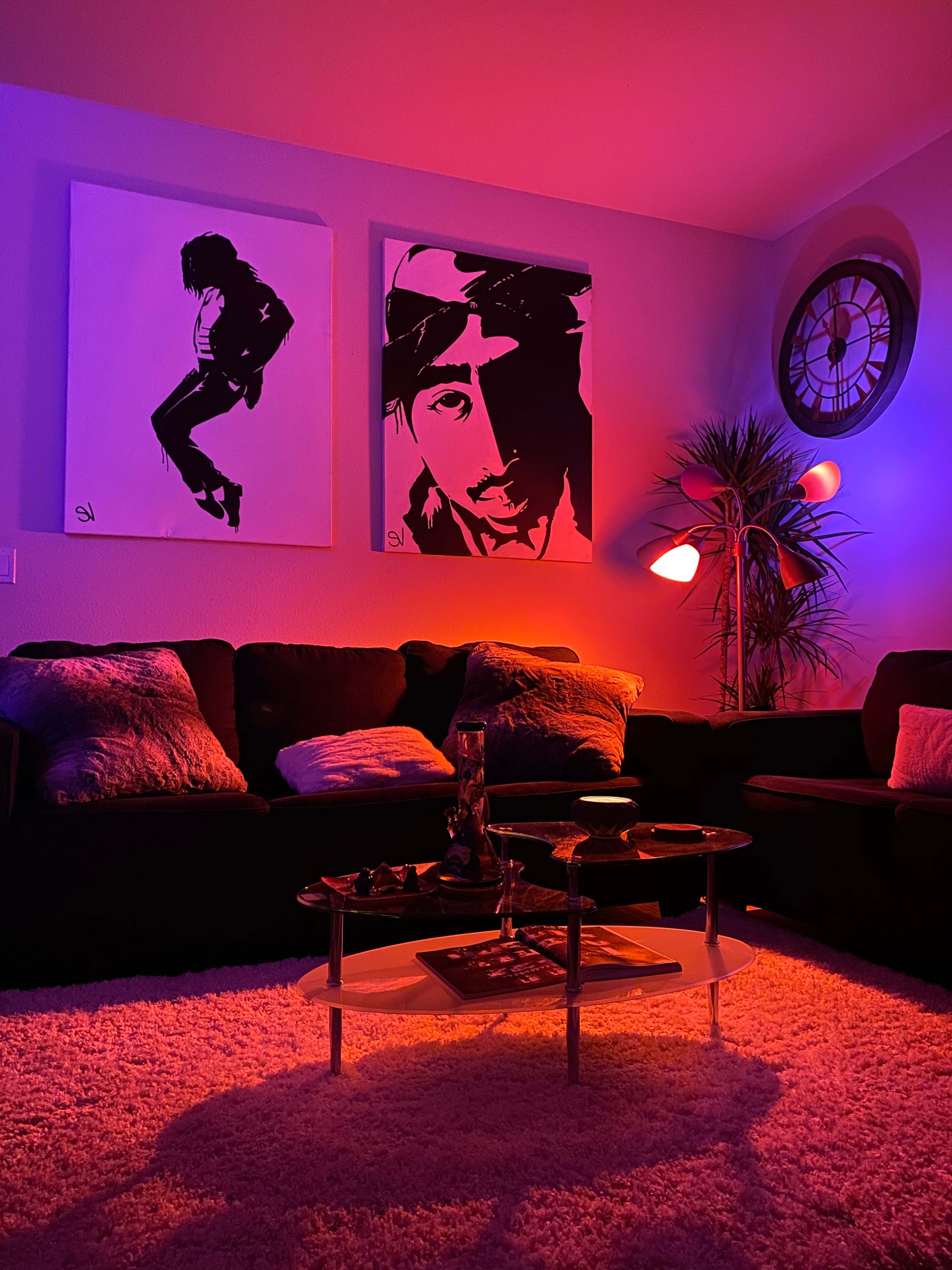 The scene features a dimly lit living room with two large framed portraits on the wall, a dark sofa with decorative pillows, and a coffee table holding glassware, all illuminated by purple and orange hues.