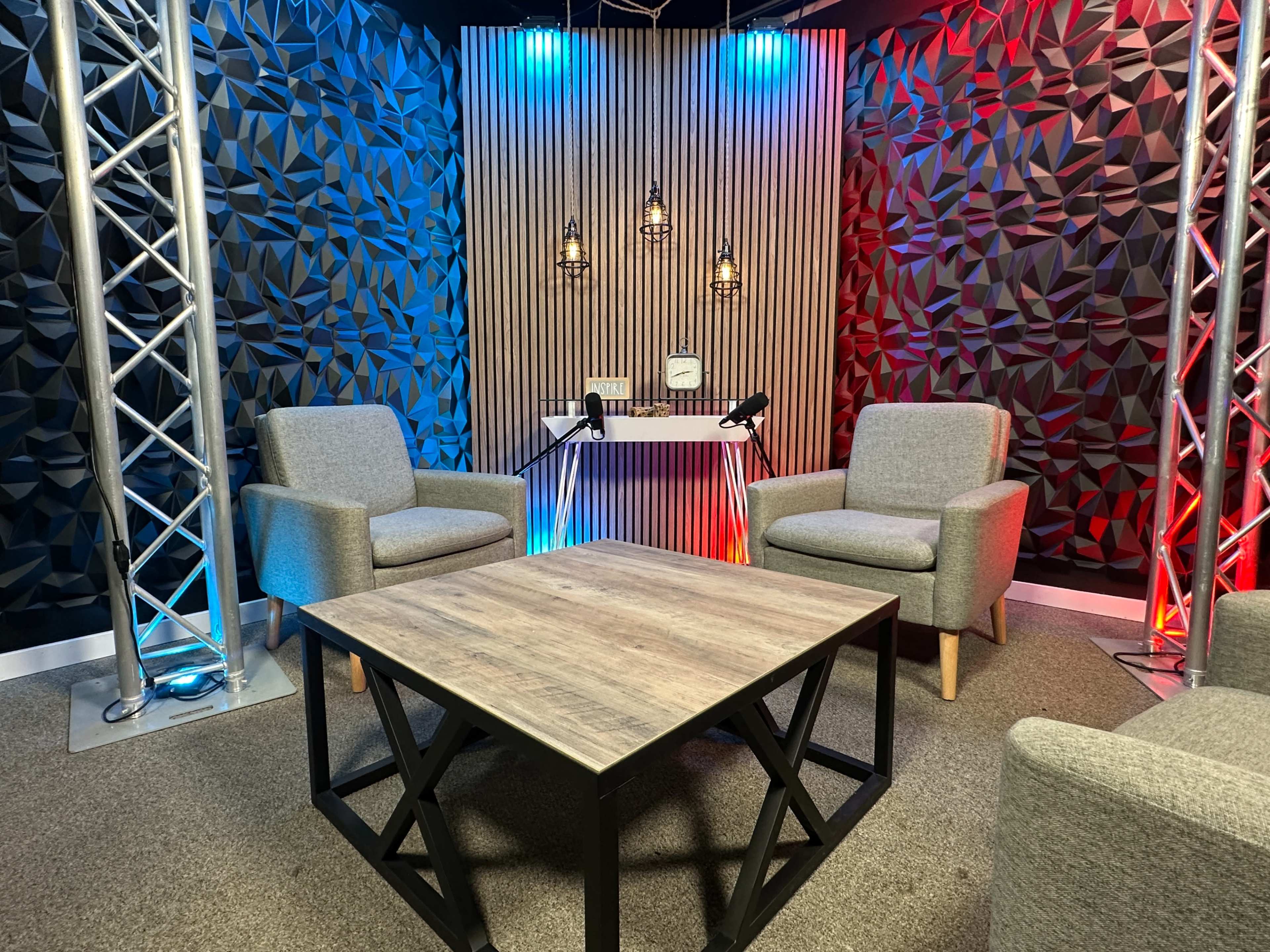 The image shows a modern studio setup with two gray armchairs and a wooden table, surrounded by textured wall panels in shades of red and blue, with decorative lighting.