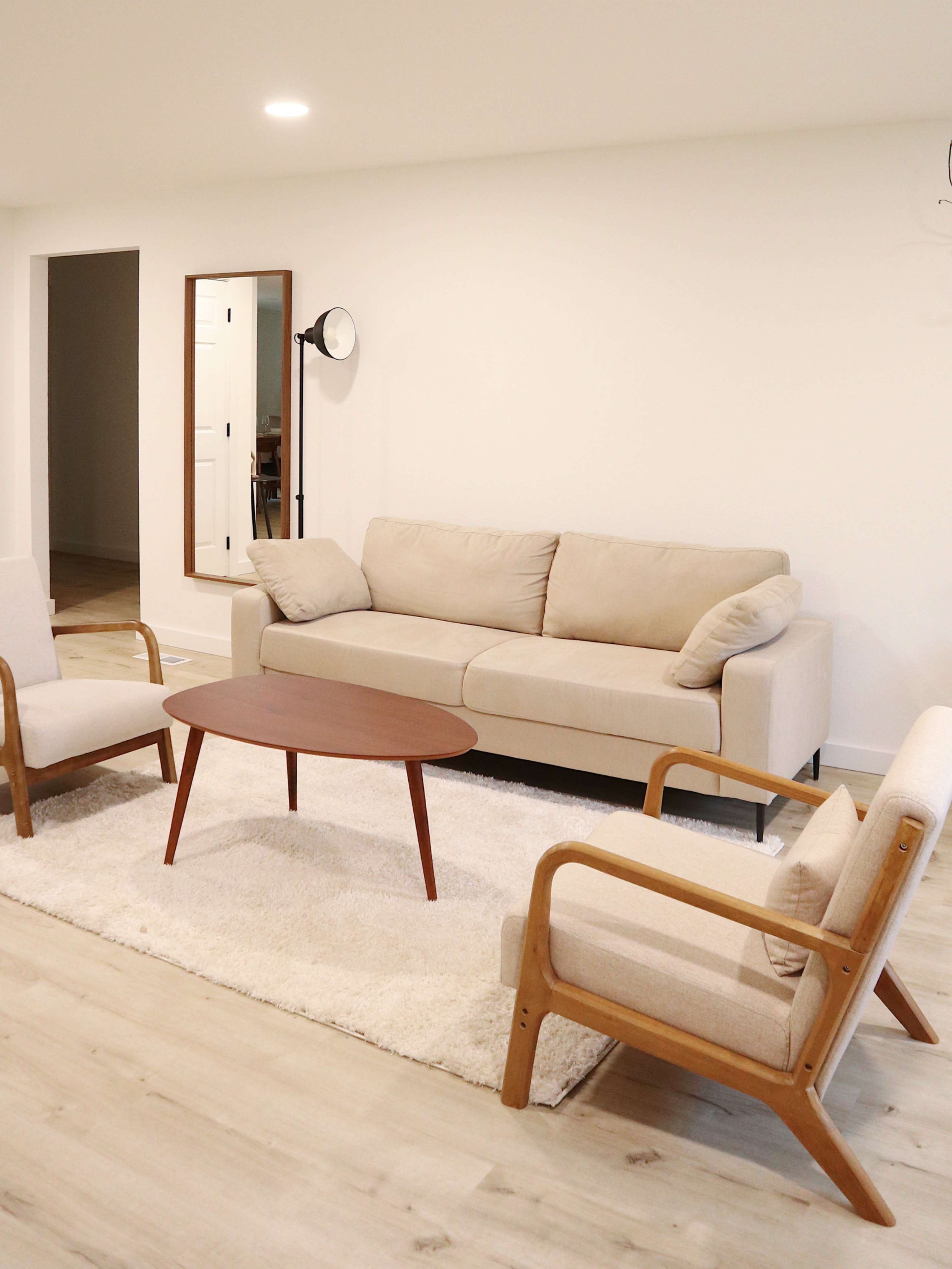 A living room features a beige sofa, two wooden armchairs, a round wooden coffee table, and a light rug.