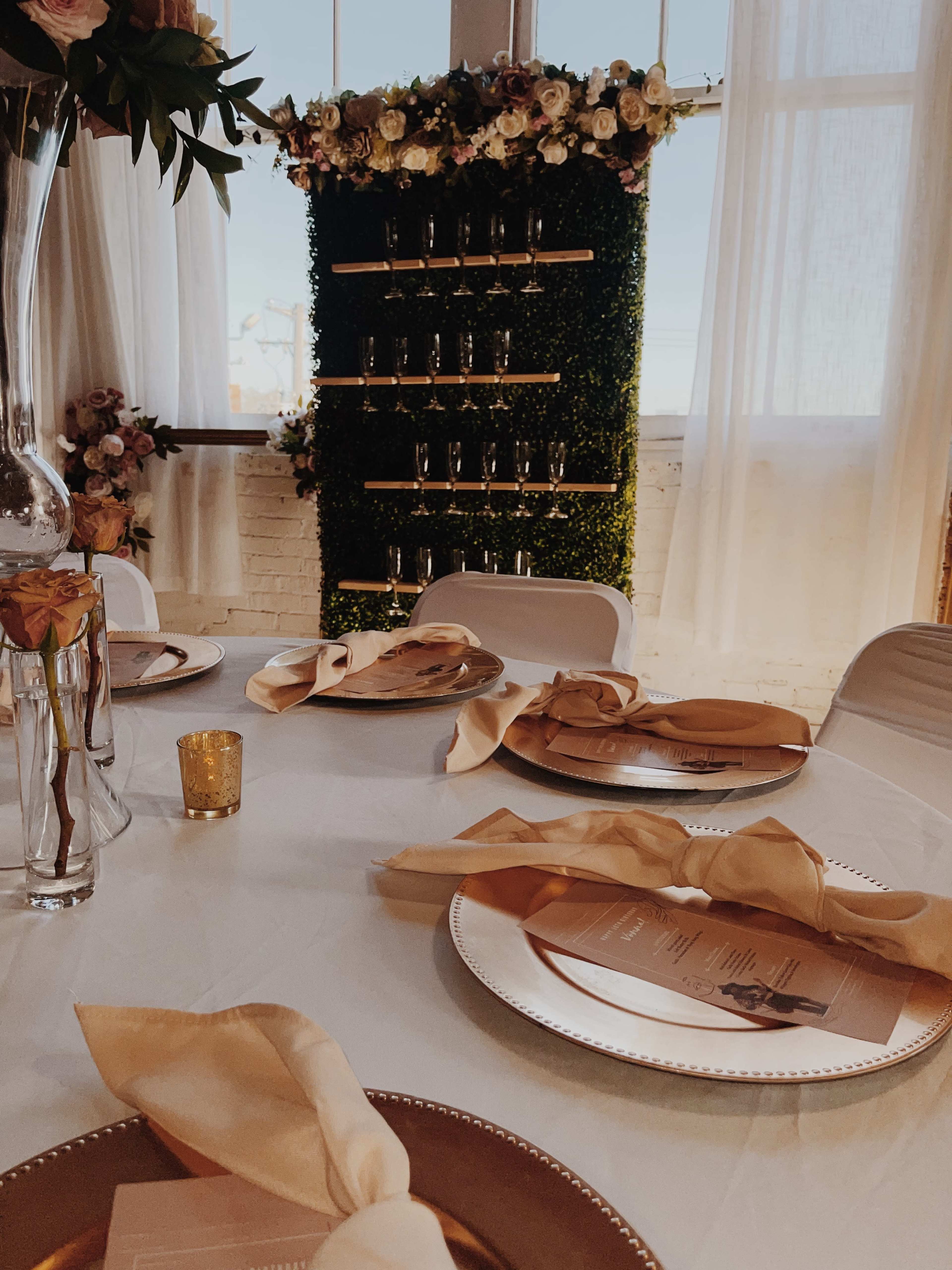 A elegantly set dining table with golden plates, napkins, and a backdrop of greenery adorned with flowers.