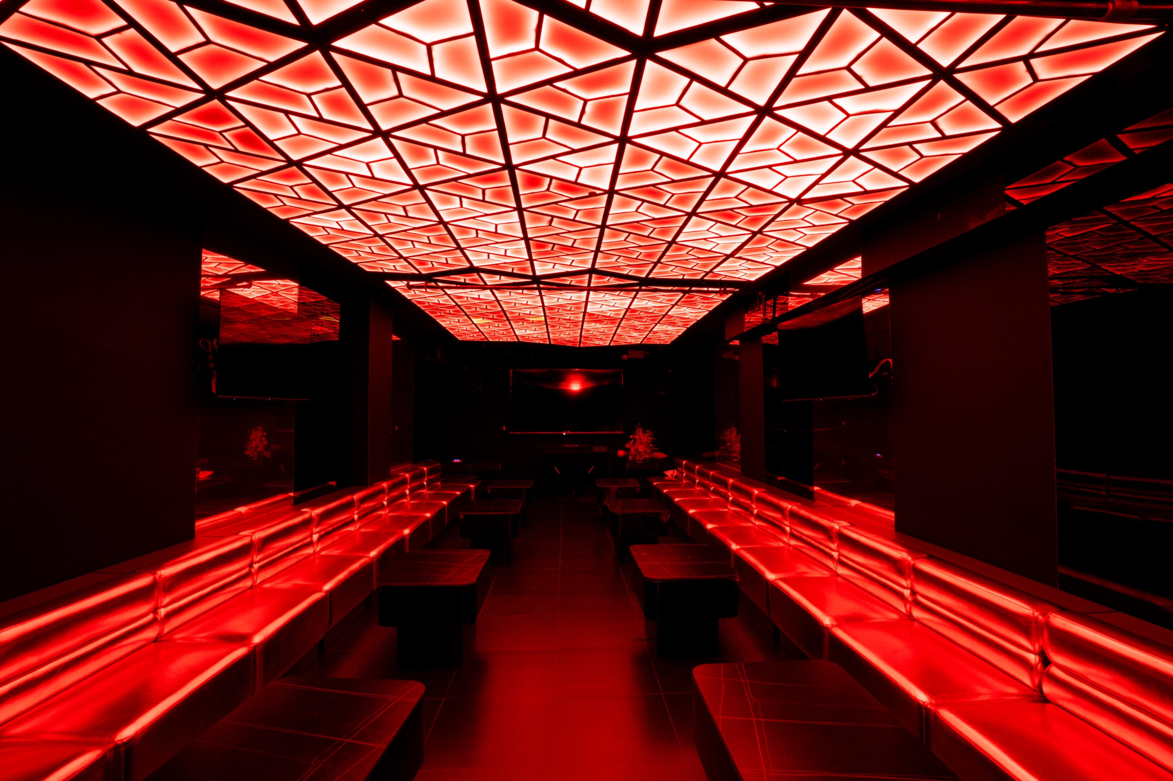The image shows a modern interior of a dimly lit lounge with red illuminated geometric ceiling tiles and black seating areas.