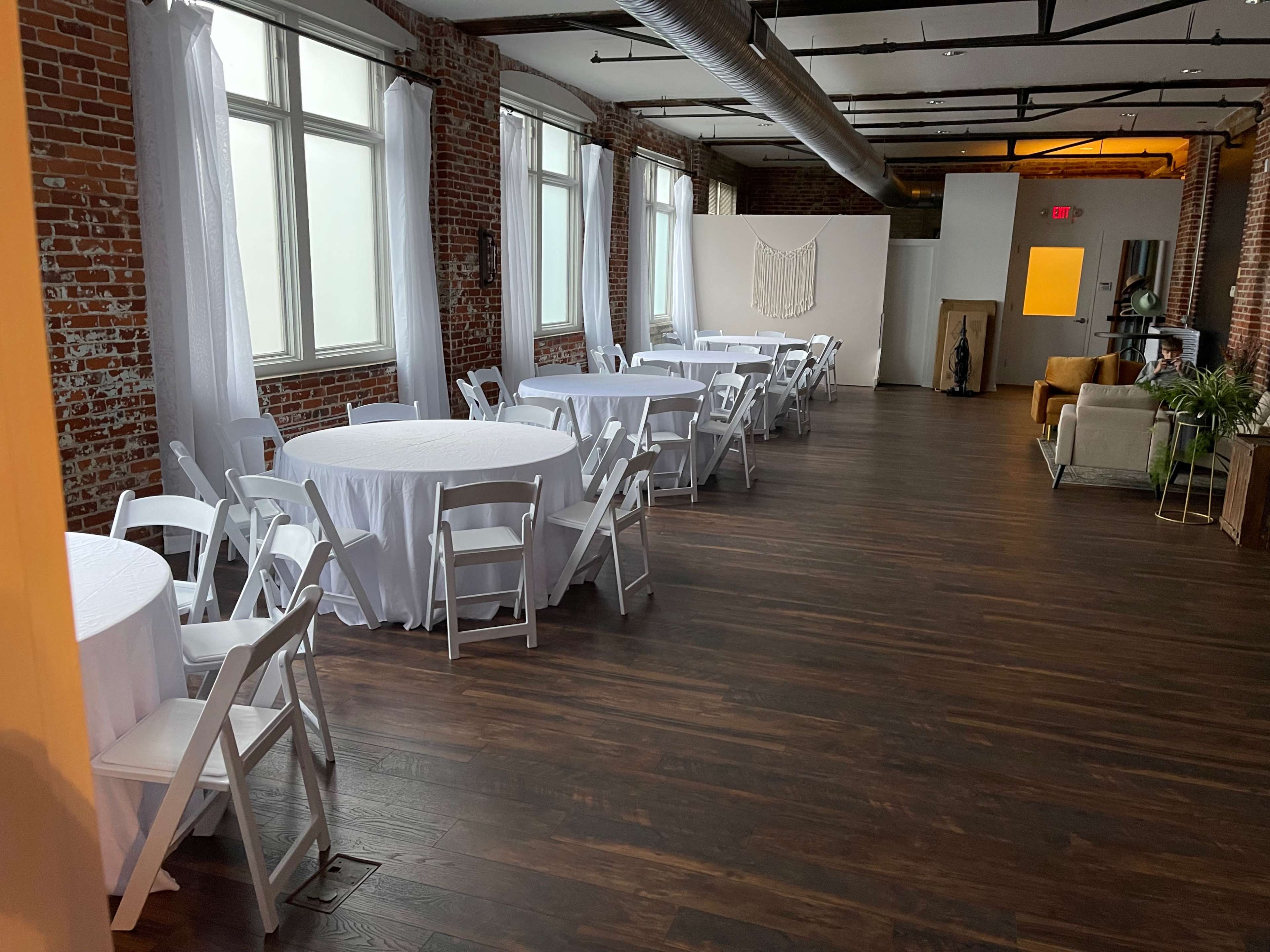 The image shows a spacious interior with white-clothed round tables arranged neatly in rows among a brick wall backdrop and large windows.