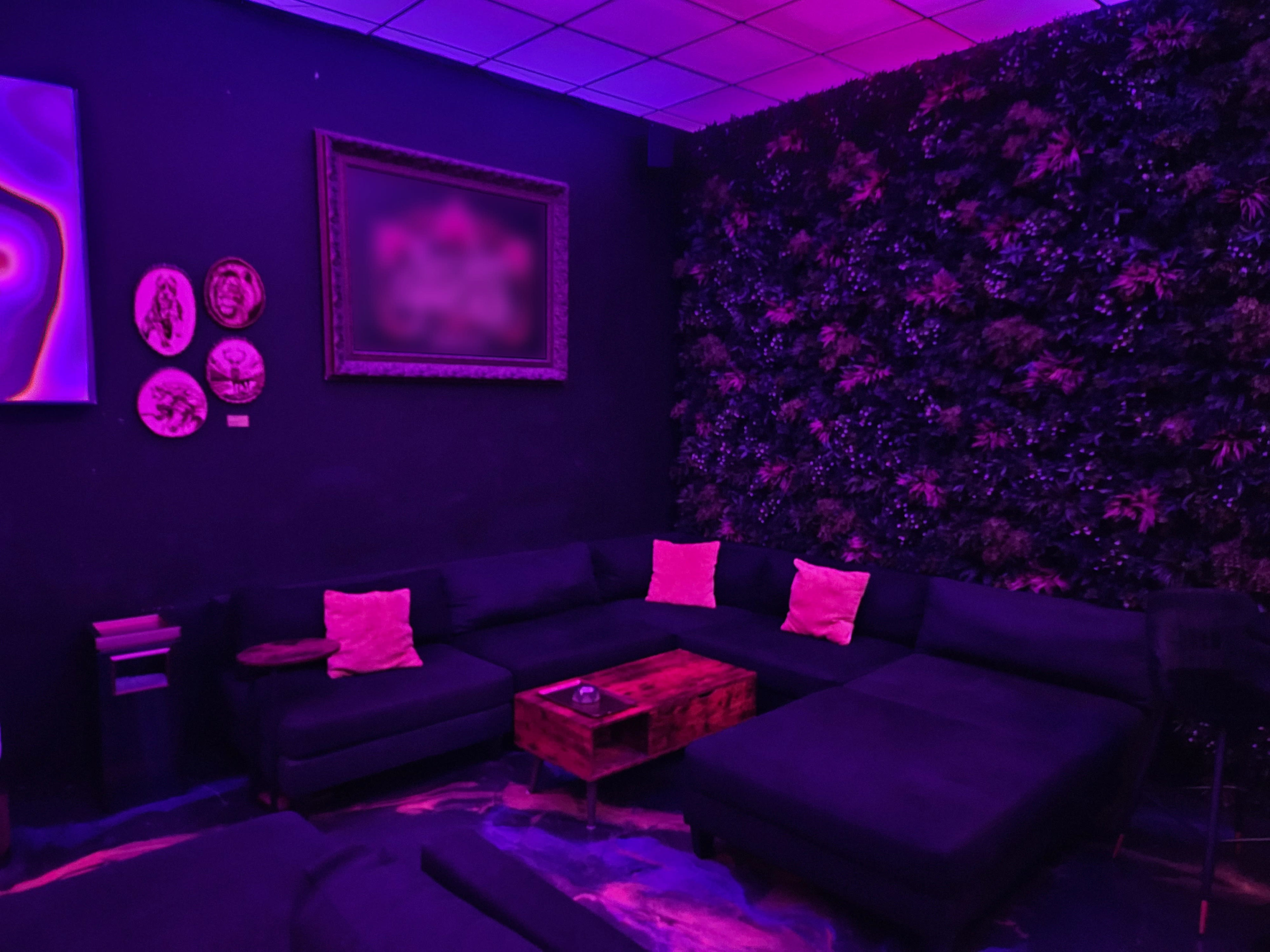 The image features a dimly lit lounge area with a purple color scheme, showcasing a sectional couch, a wooden coffee table, and a wall covered in greenery.