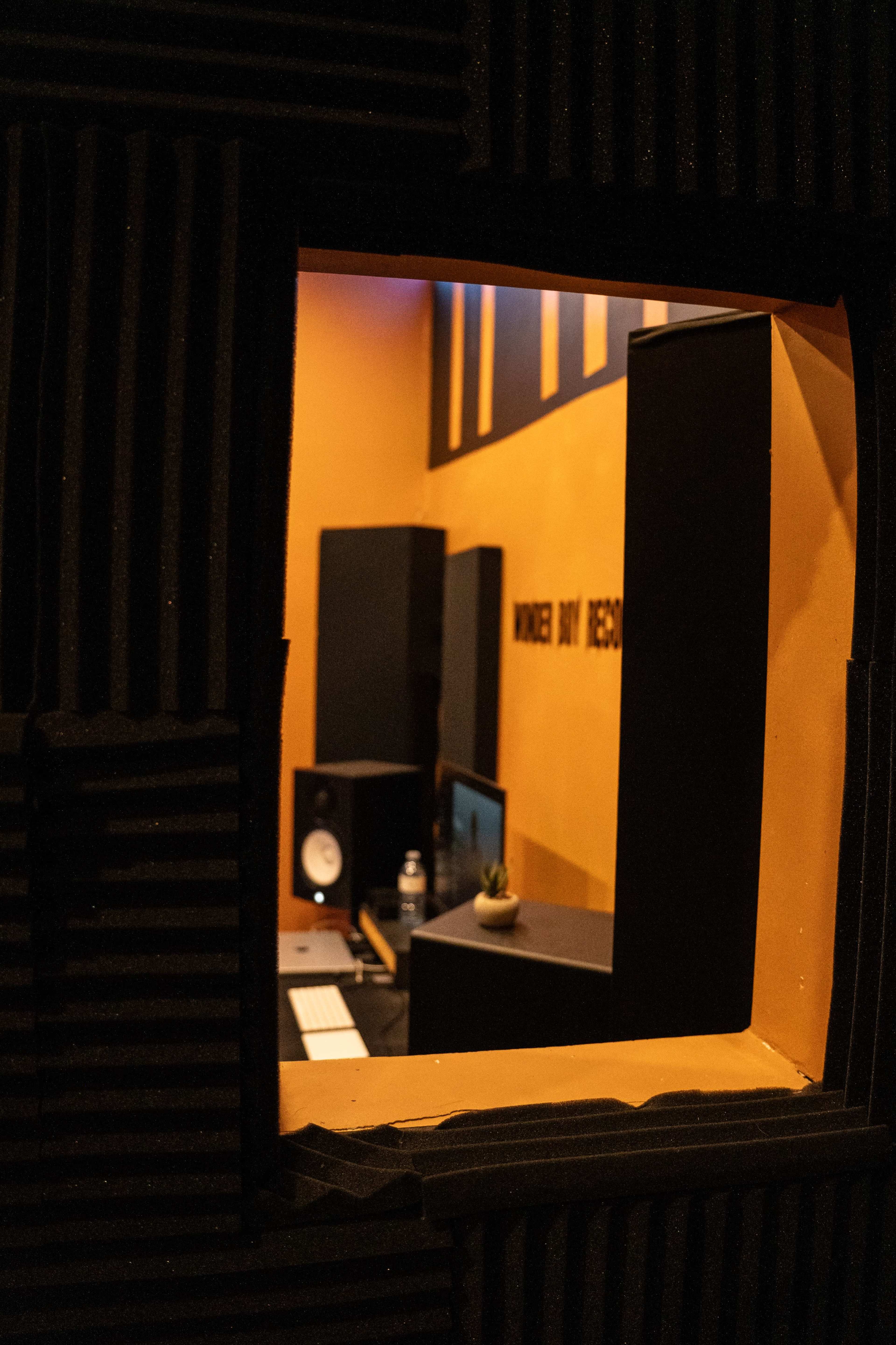 A soundproof recording studio is visible through a window, featuring black foam panels, large speakers, and a computer setup.