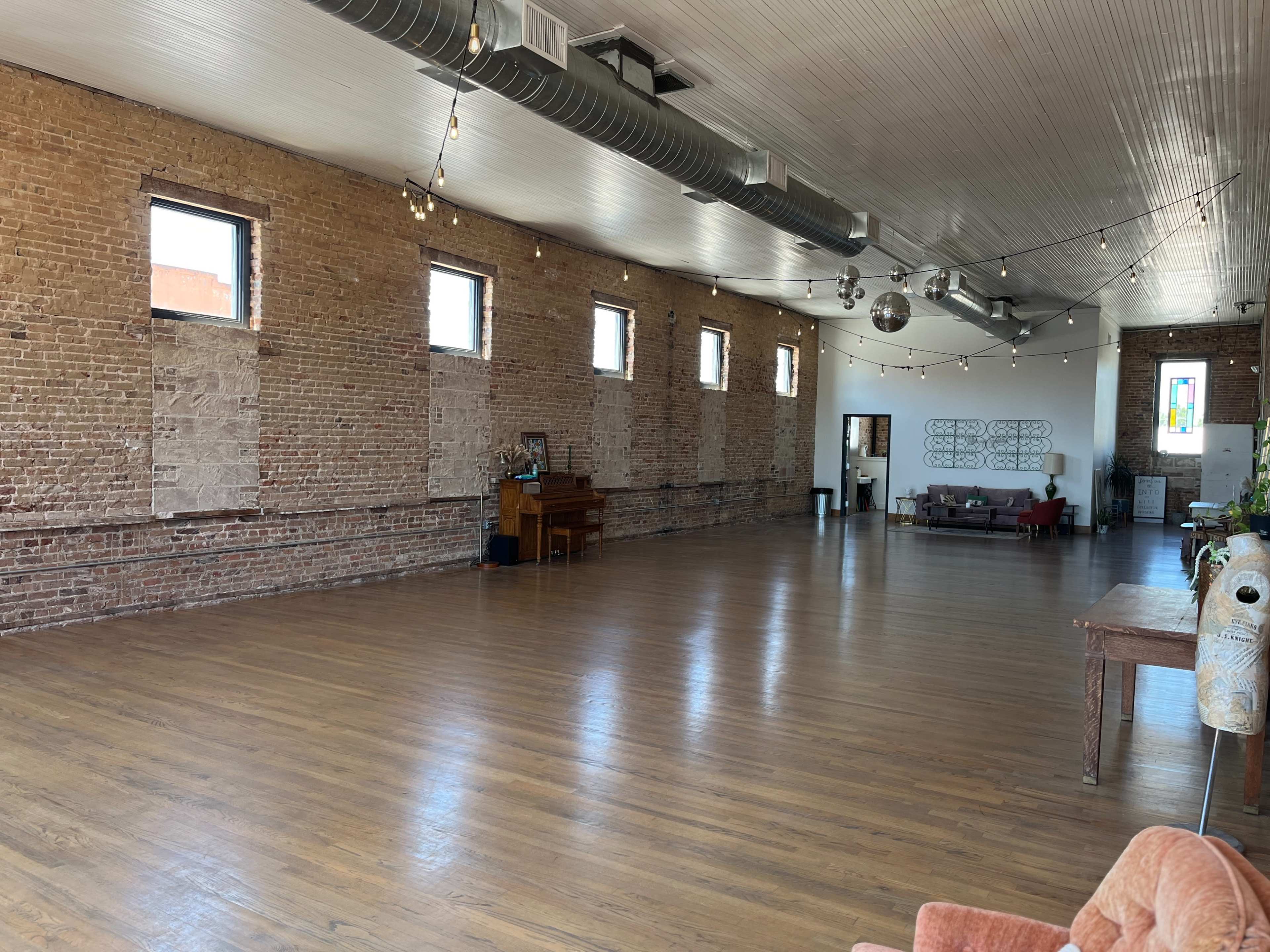 A spacious interior with exposed brick walls, hardwood floors, and large windows, featuring a piano in one corner and a seating area with a couch in another.