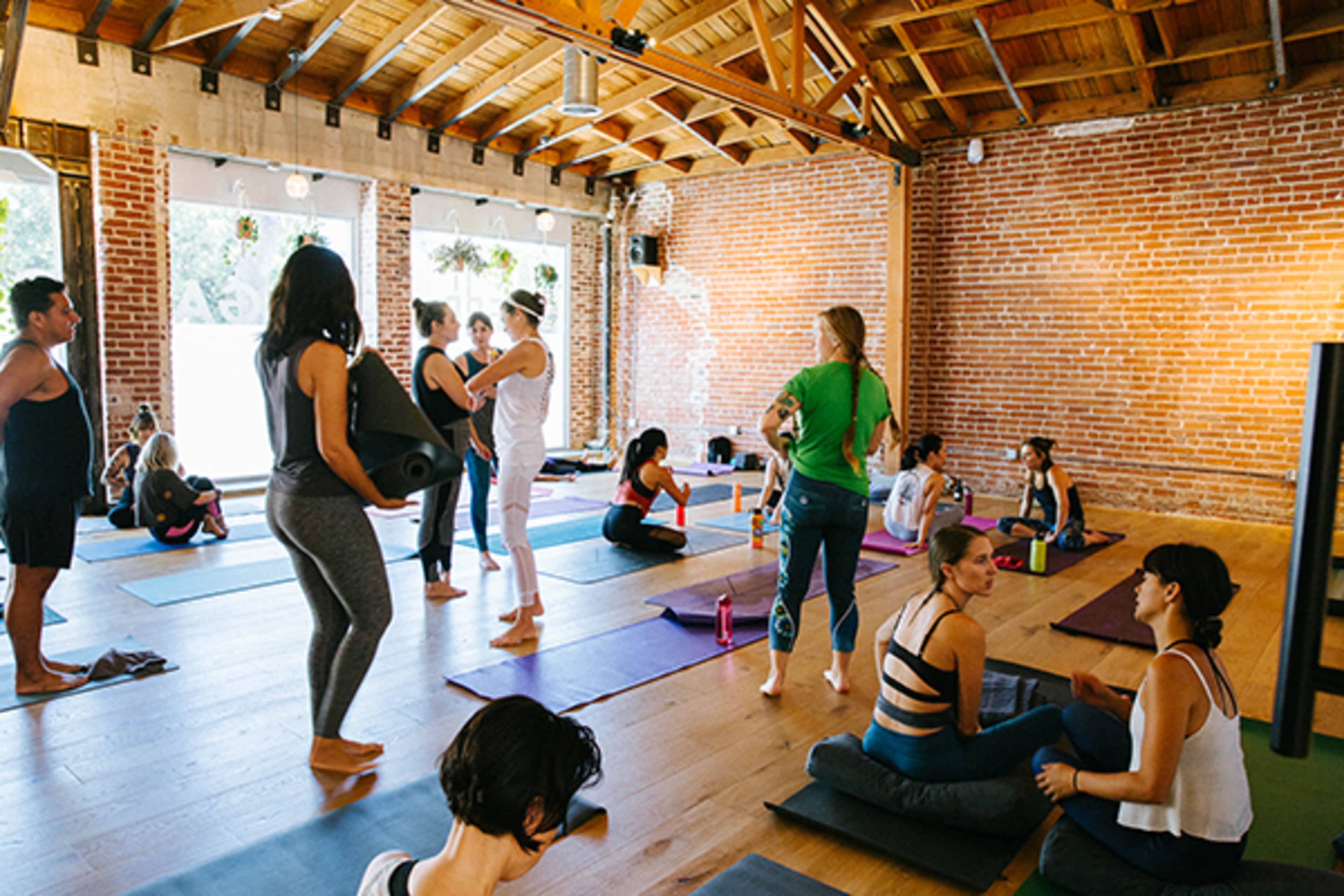 A yoga class is in session in a spacious studio with wooden floors and brick walls, where participants are engaged in various activities, including stretching, talking, and preparing their mats.
