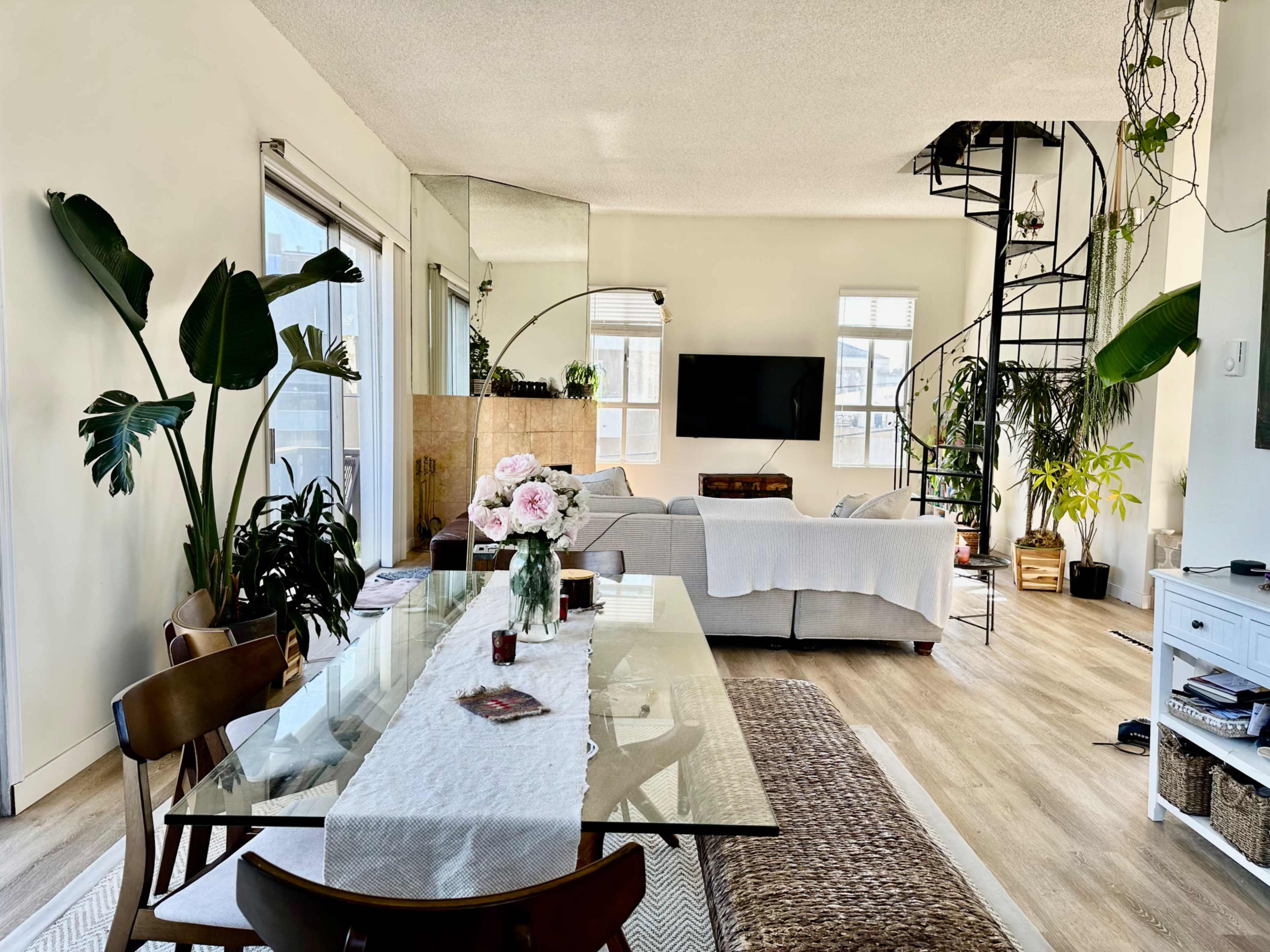 A bright, open living space features a dining area with a glass table and plants, a cozy seating area with a television, and a spiral staircase leading to an upper level.