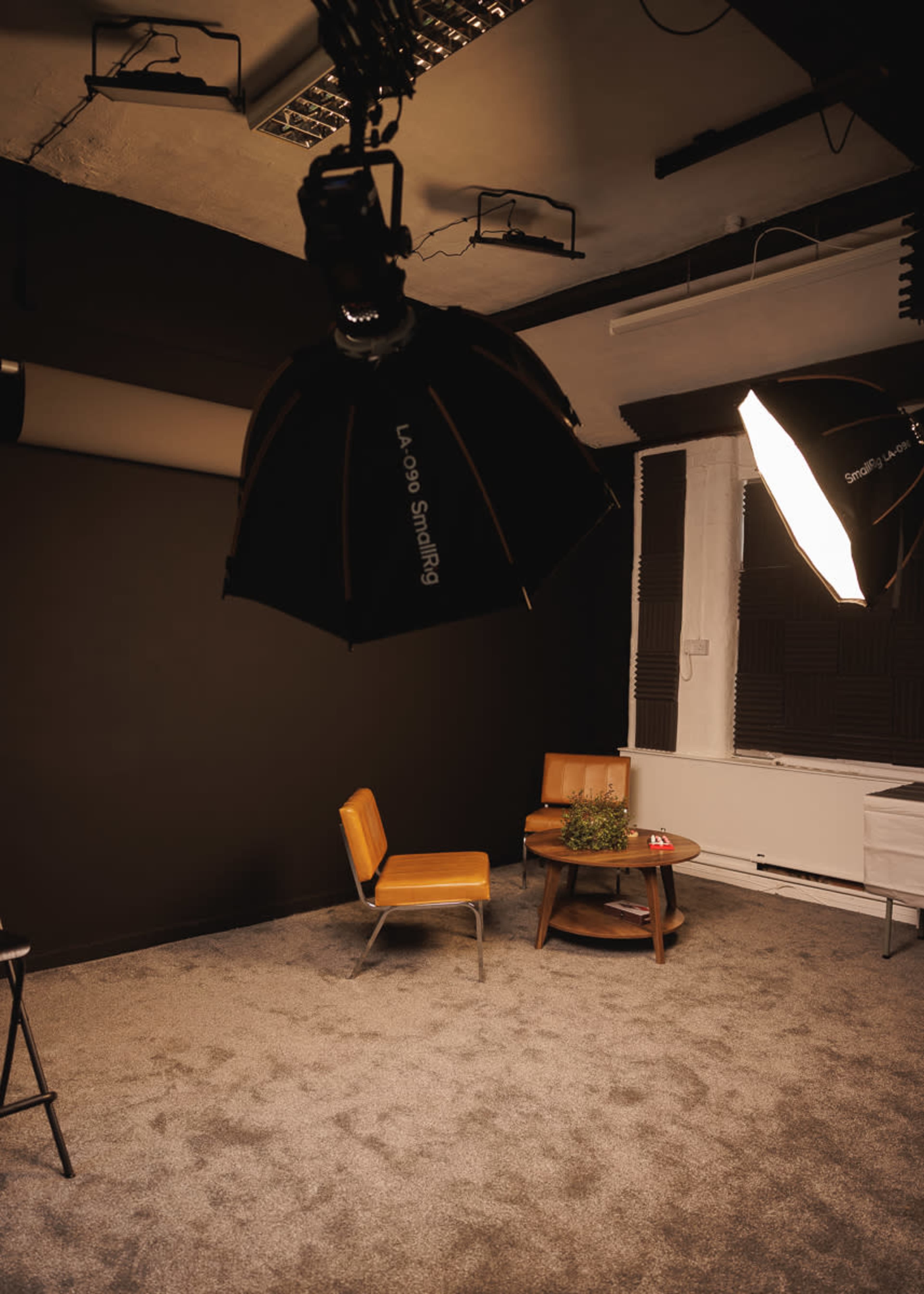 The image shows a studio room featuring a brown wall, a circular table with a plant, and a single orange chair under studio lighting.