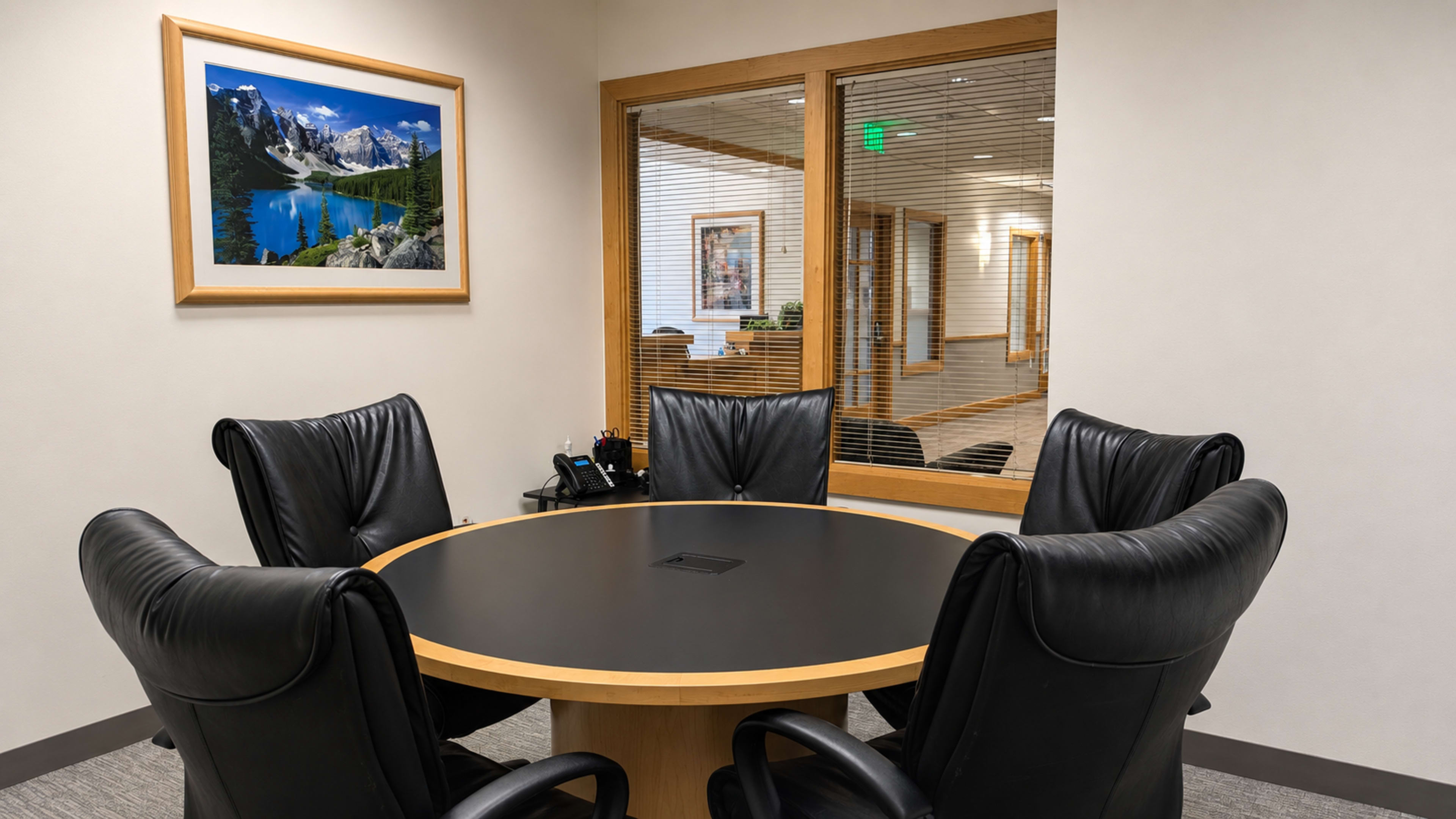 The image shows a conference room with a round table surrounded by four black leather chairs and a window overlooking a hallway.