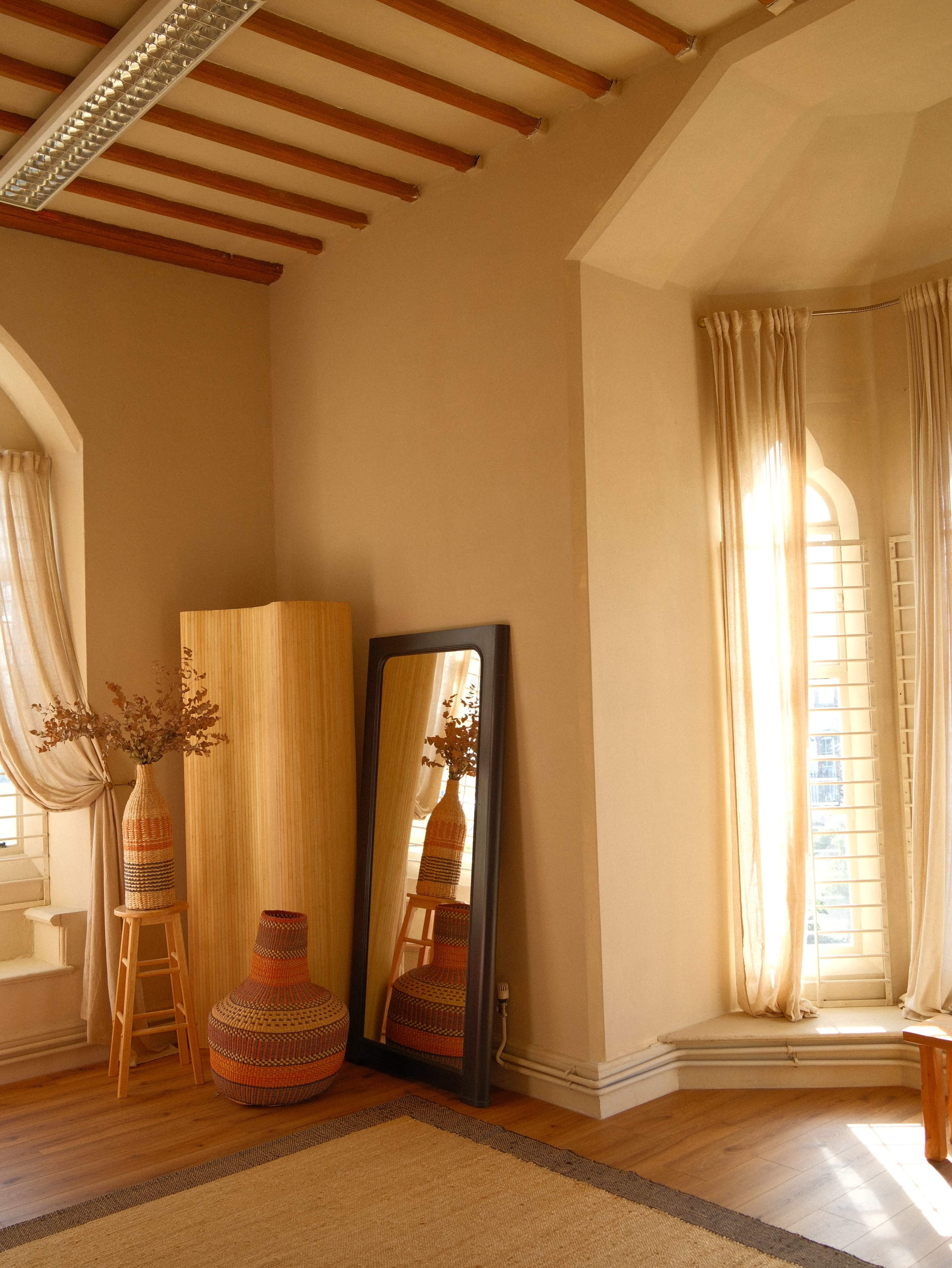 The image shows a sunlit room with a tall mirror reflecting a corner featuring a wooden cabinet, a decorative vase on a stool, and soft curtains framing arched windows.
