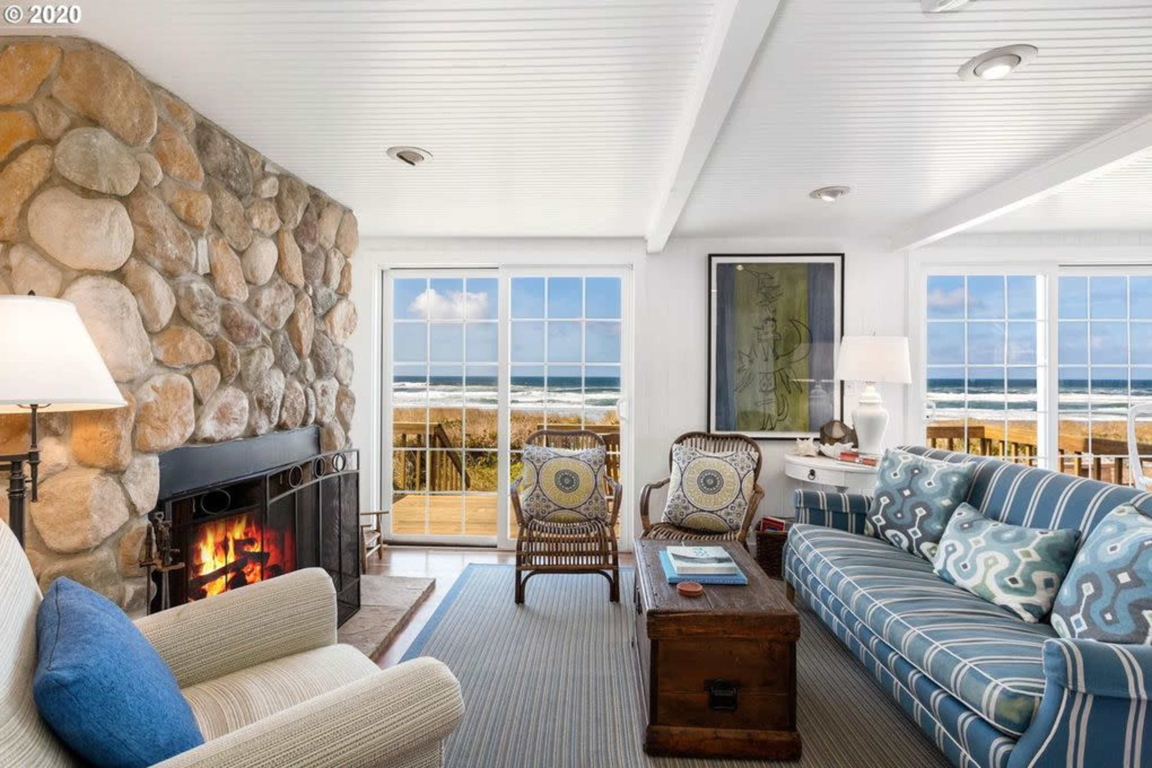 A living room features a stone fireplace, striped blue and white sofas, and large windows that open to a view of the ocean.