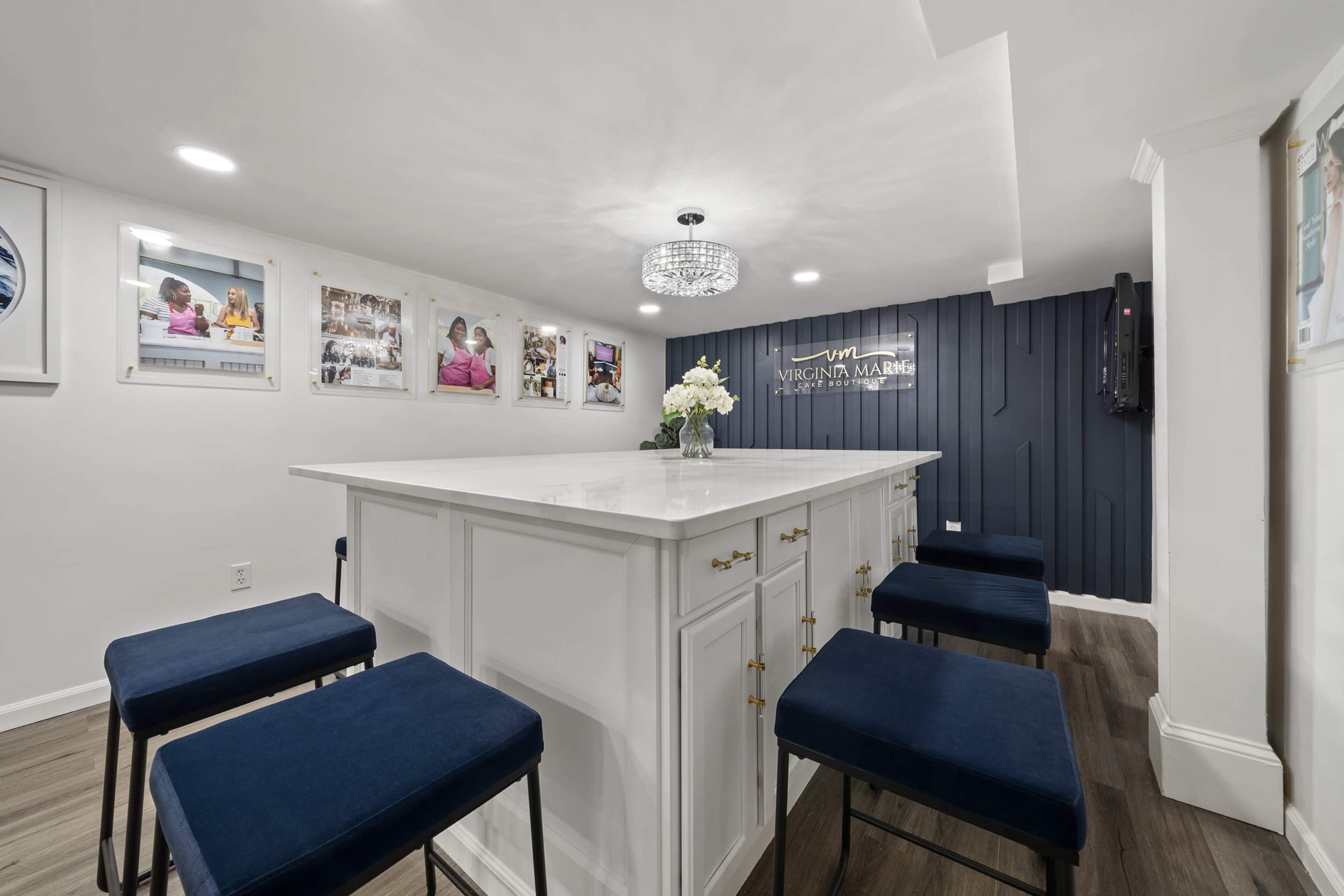 The image shows a modern interior space featuring a white kitchen island with high-backed blue stools, accented by a decorative chandelier and framed photographs on the walls.