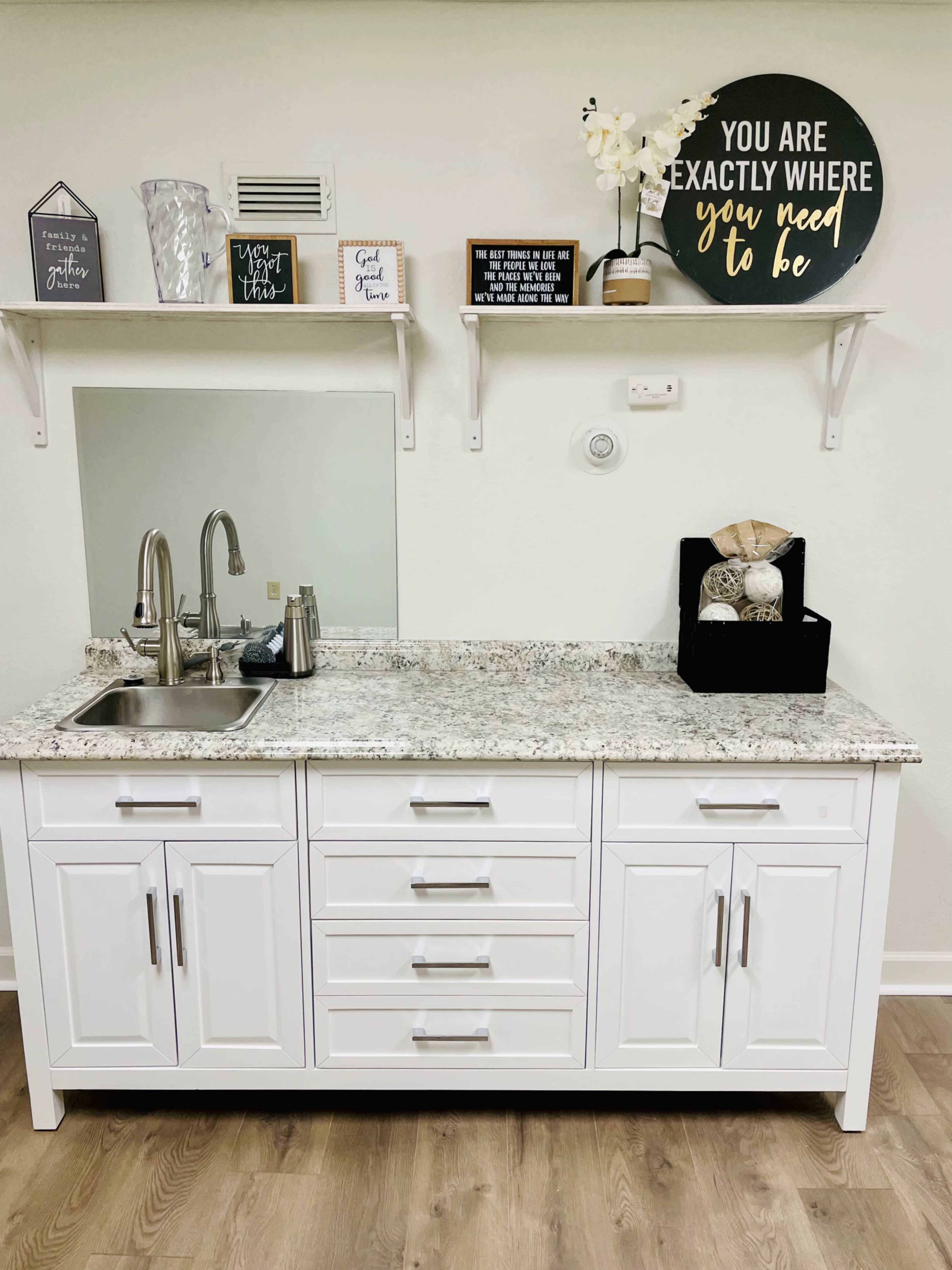 A modern white kitchen cabinet with a granite countertop, featuring a sink, decorative items on the wall shelf, and a black box on the counter.