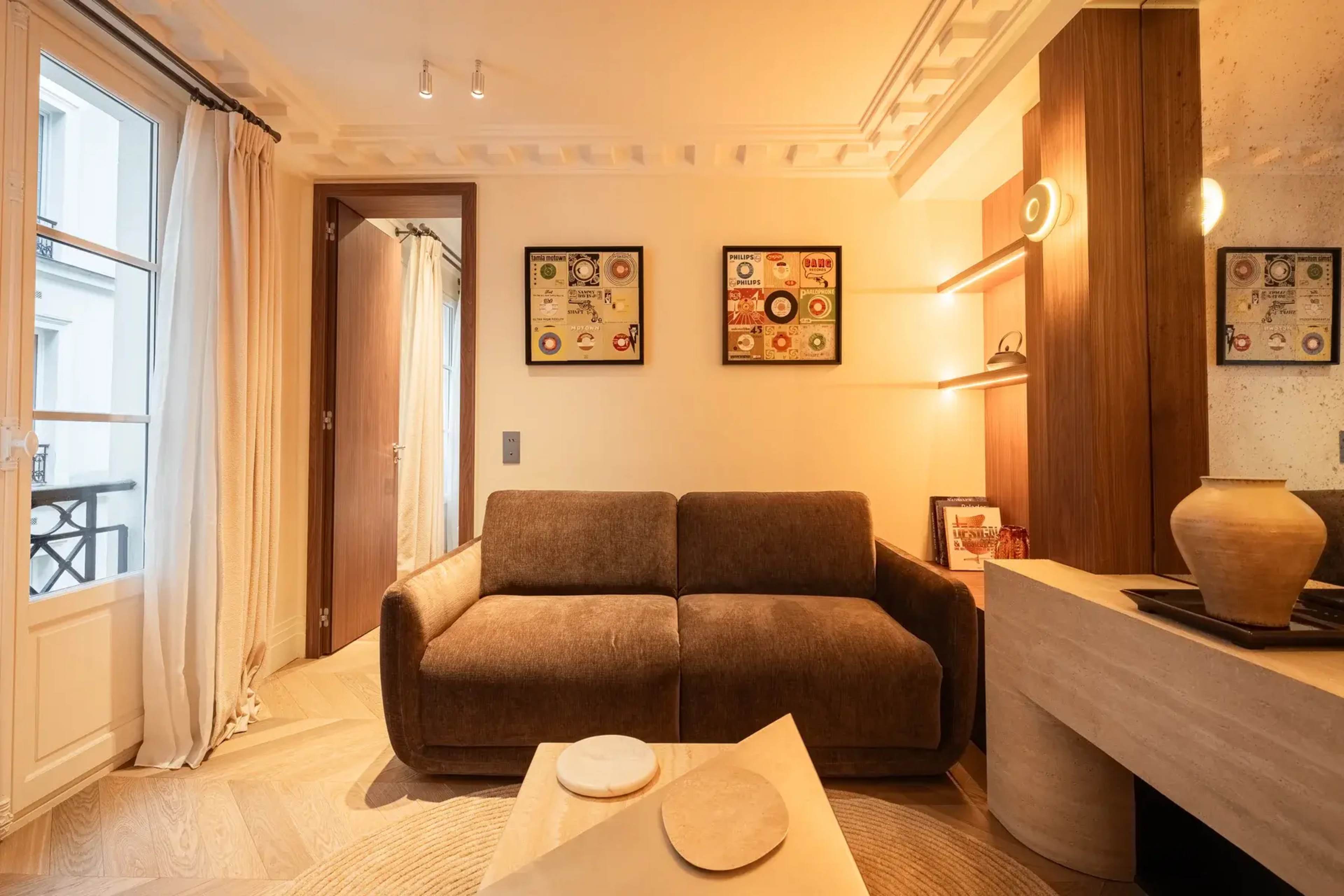 The image shows a cozy living room with a brown sofa, a decorative table, and framed artwork on the walls, illuminated by warm lighting.