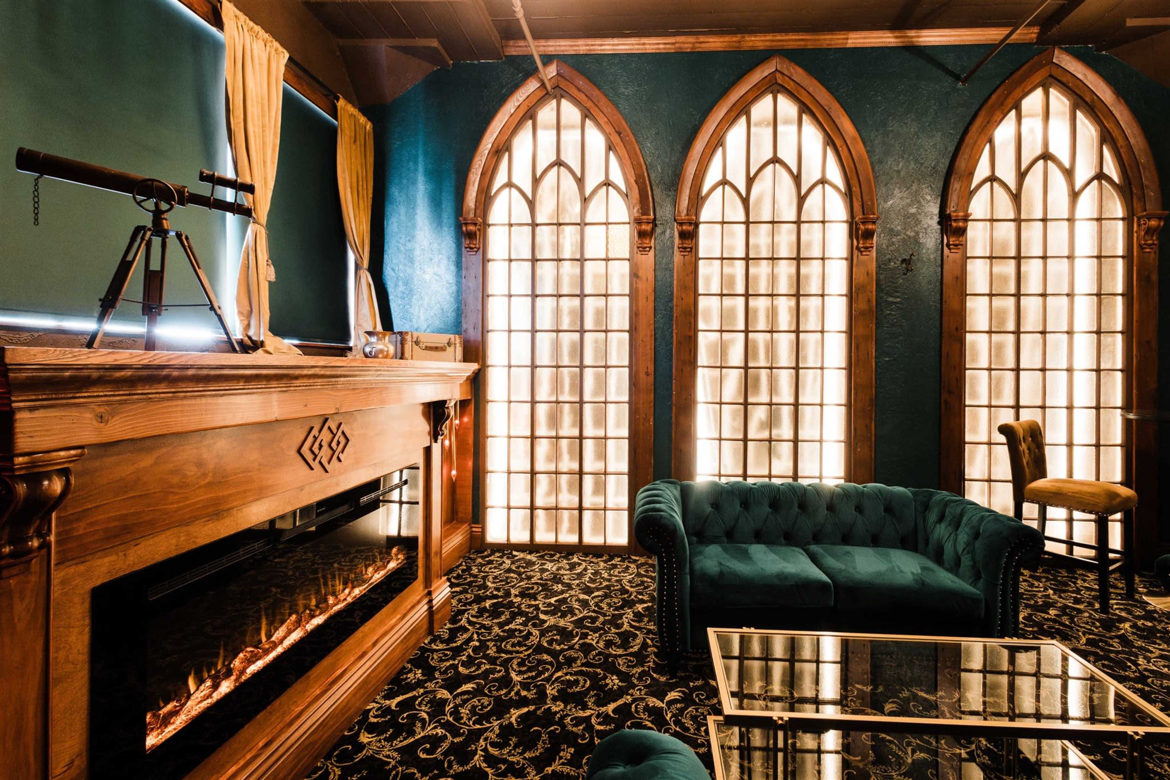 The image shows an elegantly designed room featuring a plush teal couch, a wooden fireplace with an ornate mantel, and large, arched windows that allow bright light to fill the space.