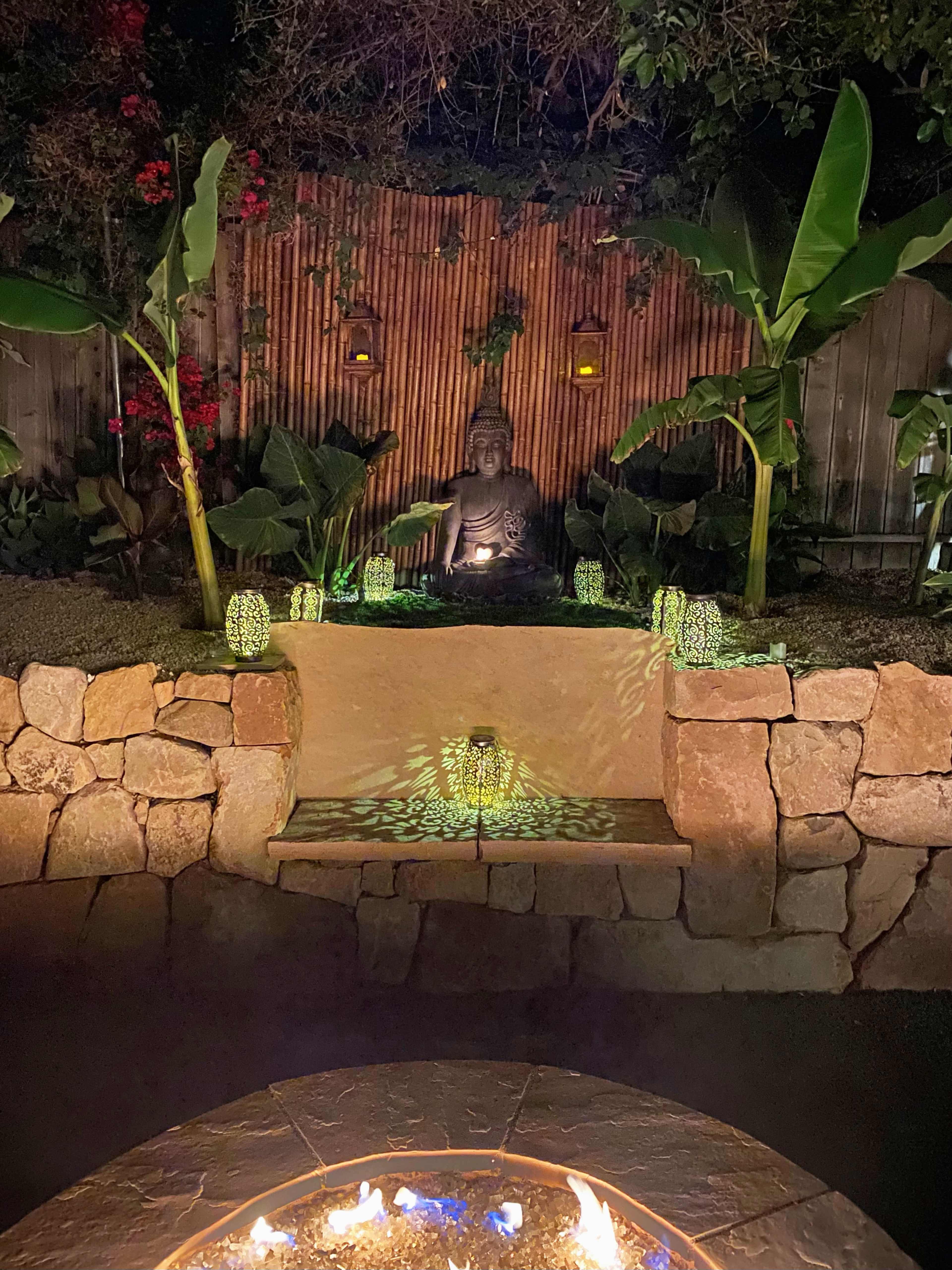 A stone platform features a seated Buddha surrounded by tropical plants, with decorative lights casting patterns on the surface near a fire pit.