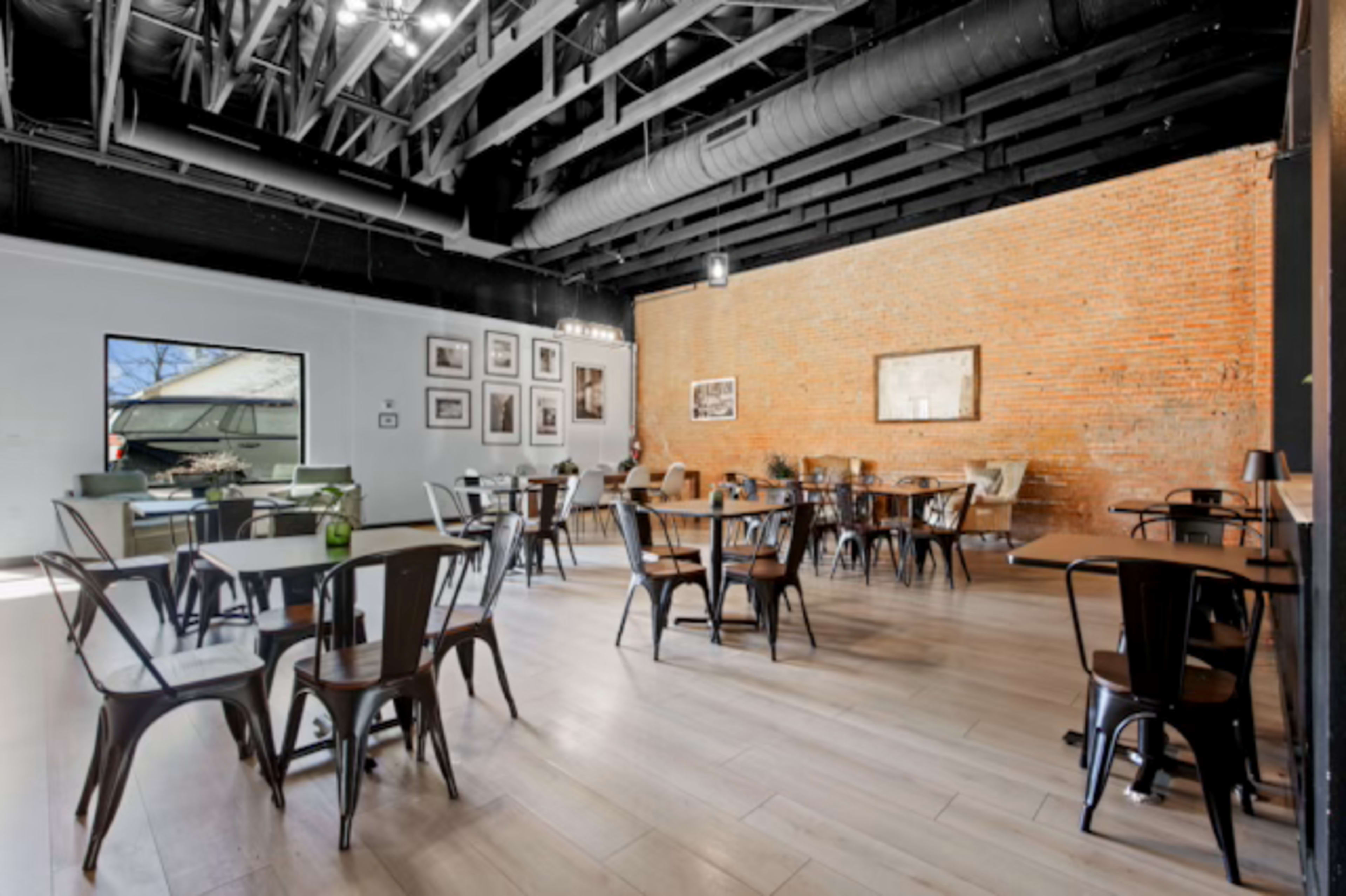 The image shows a spacious café with metal chairs and wooden tables, featuring exposed brick walls and large windows.