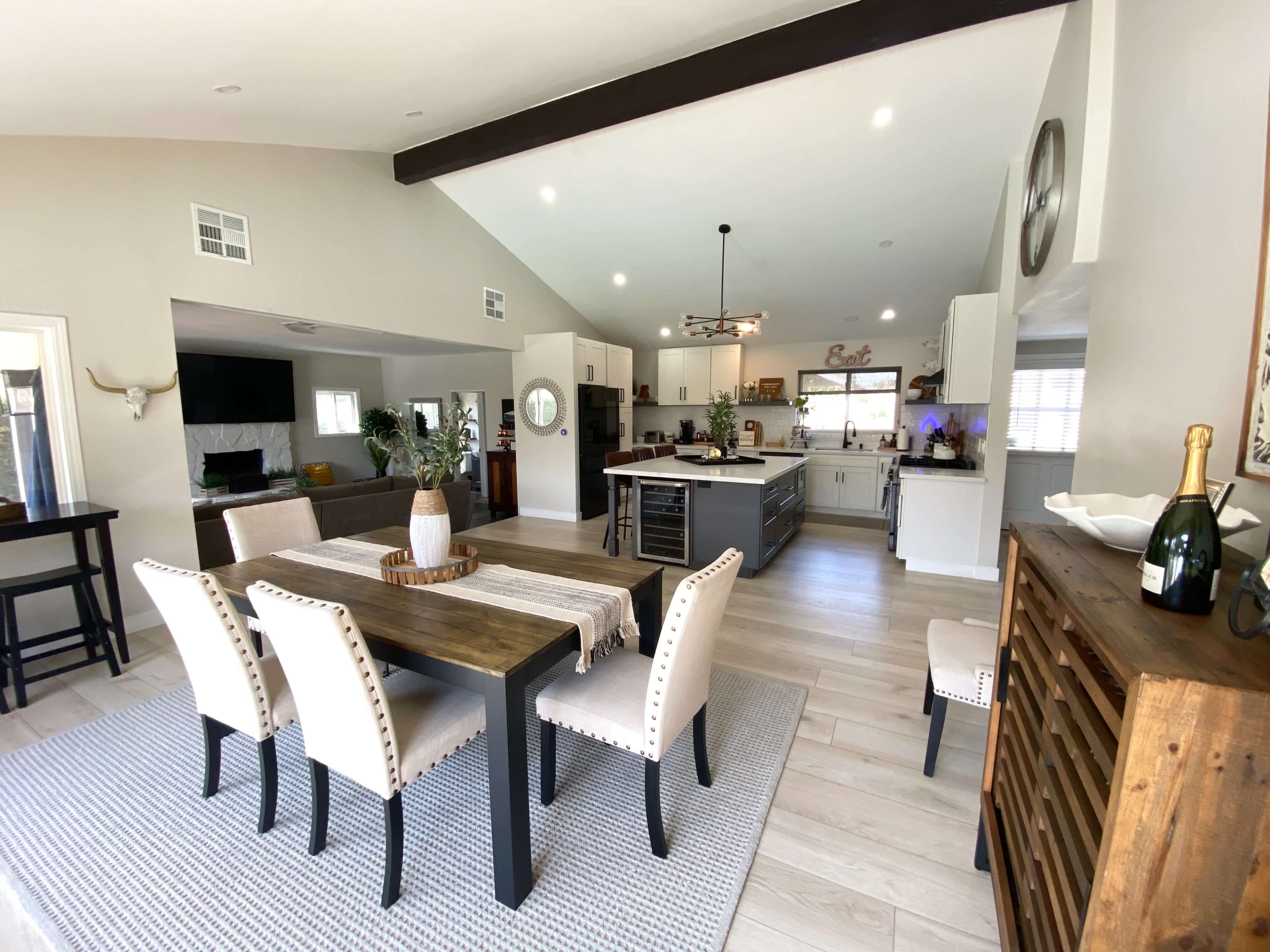 The image shows a modern, open-concept kitchen and dining area featuring a wooden dining table, beige upholstered chairs, and a kitchen island with bar seating.