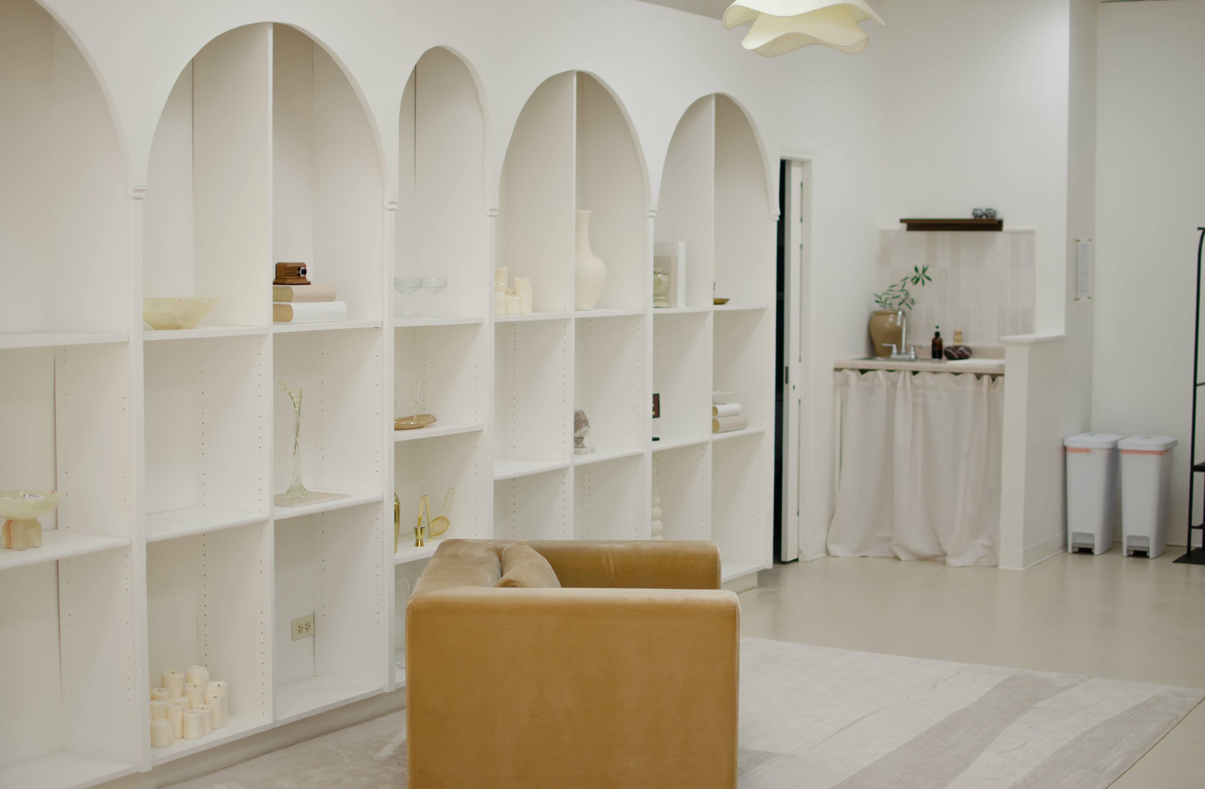 The scene features a minimalist interior with white shelves in arch shapes, a tan sofa, and a small kitchen area with a curtain.