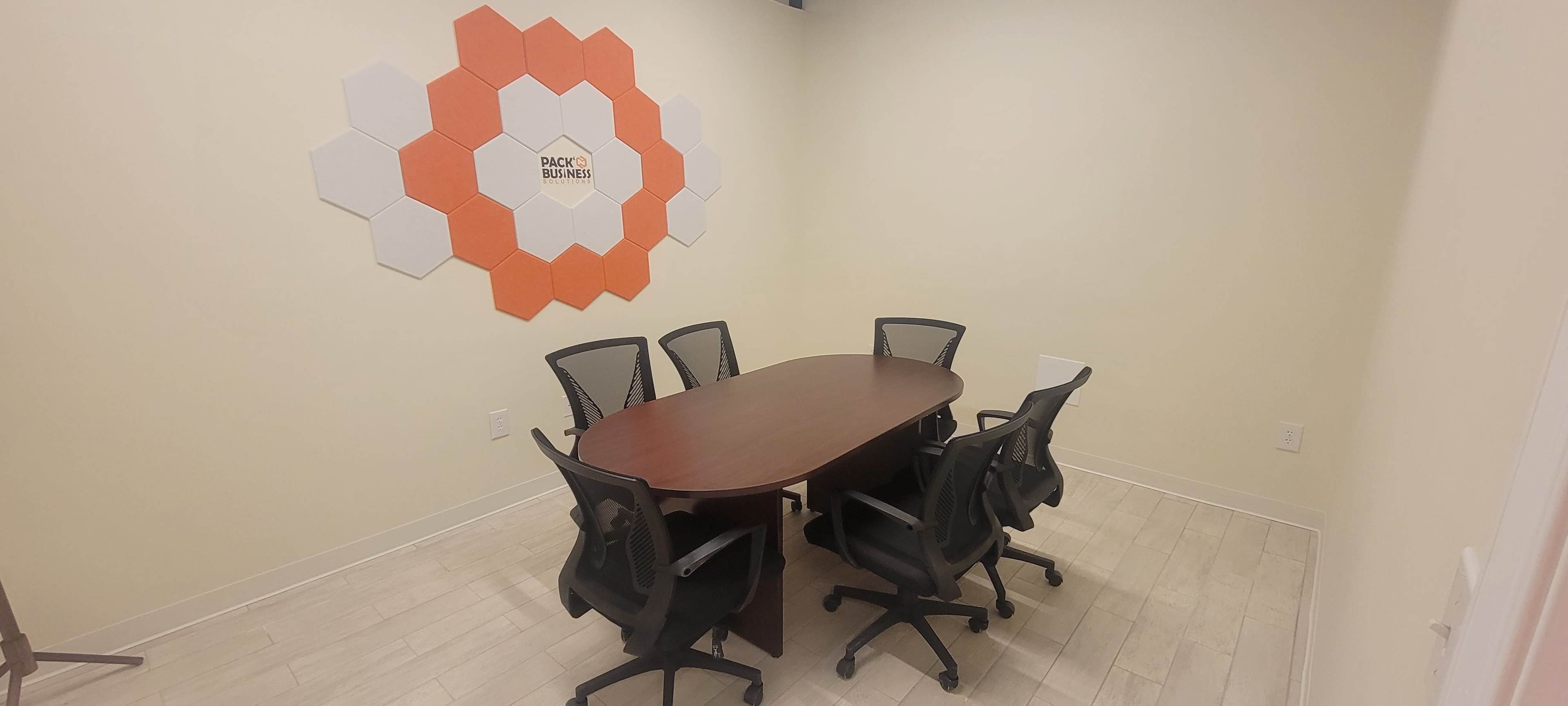 A meeting room features a wooden oval table surrounded by six ergonomic chairs, with a hexagonal wall design in orange and white.