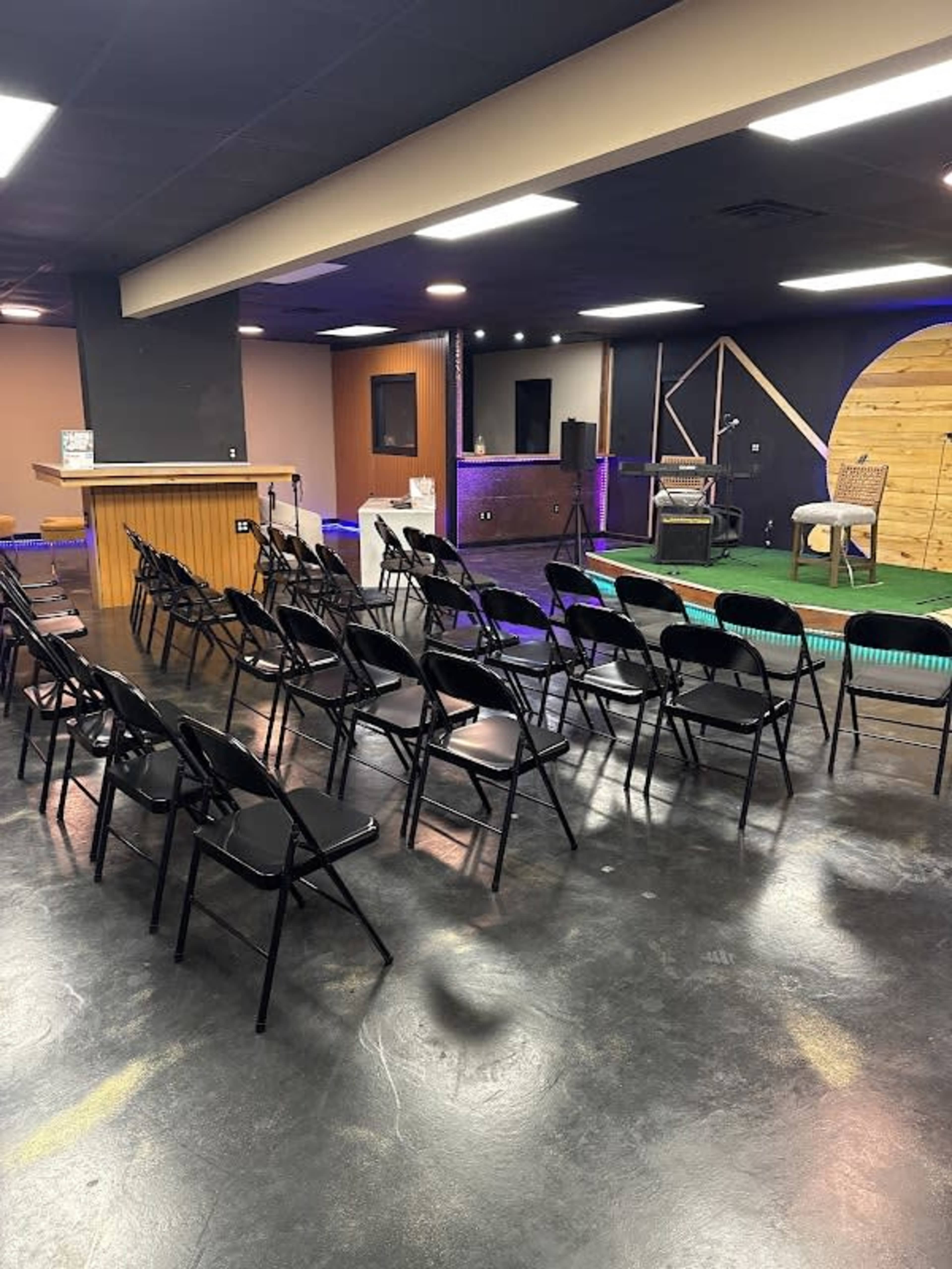 The image shows a room arranged for an event, featuring rows of black chairs facing a stage area with a small podium and a green carpet.