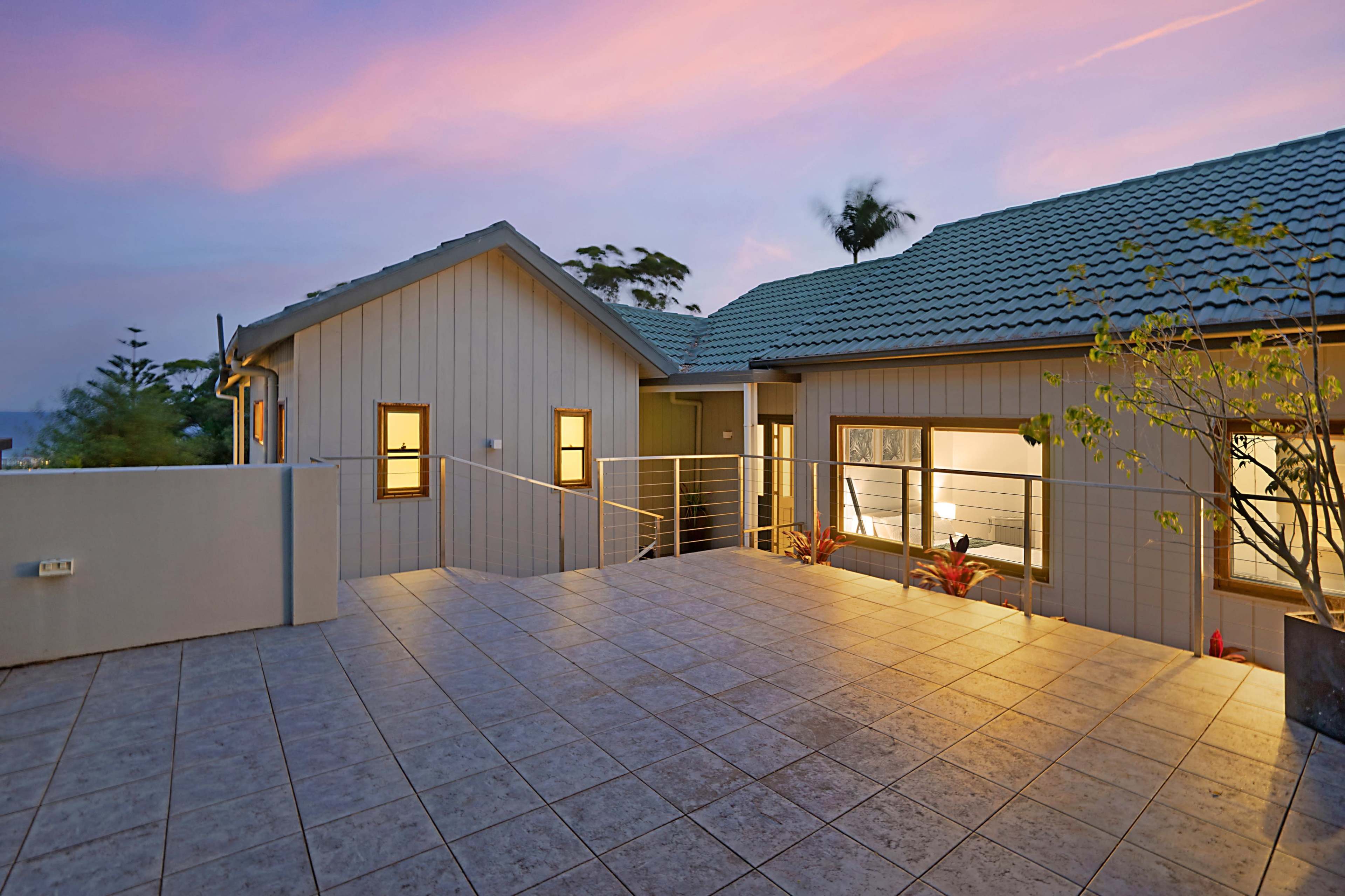 A modern house features a spacious patio with tile flooring, surrounded by landscaping and framed by a colorful sky at sunset.